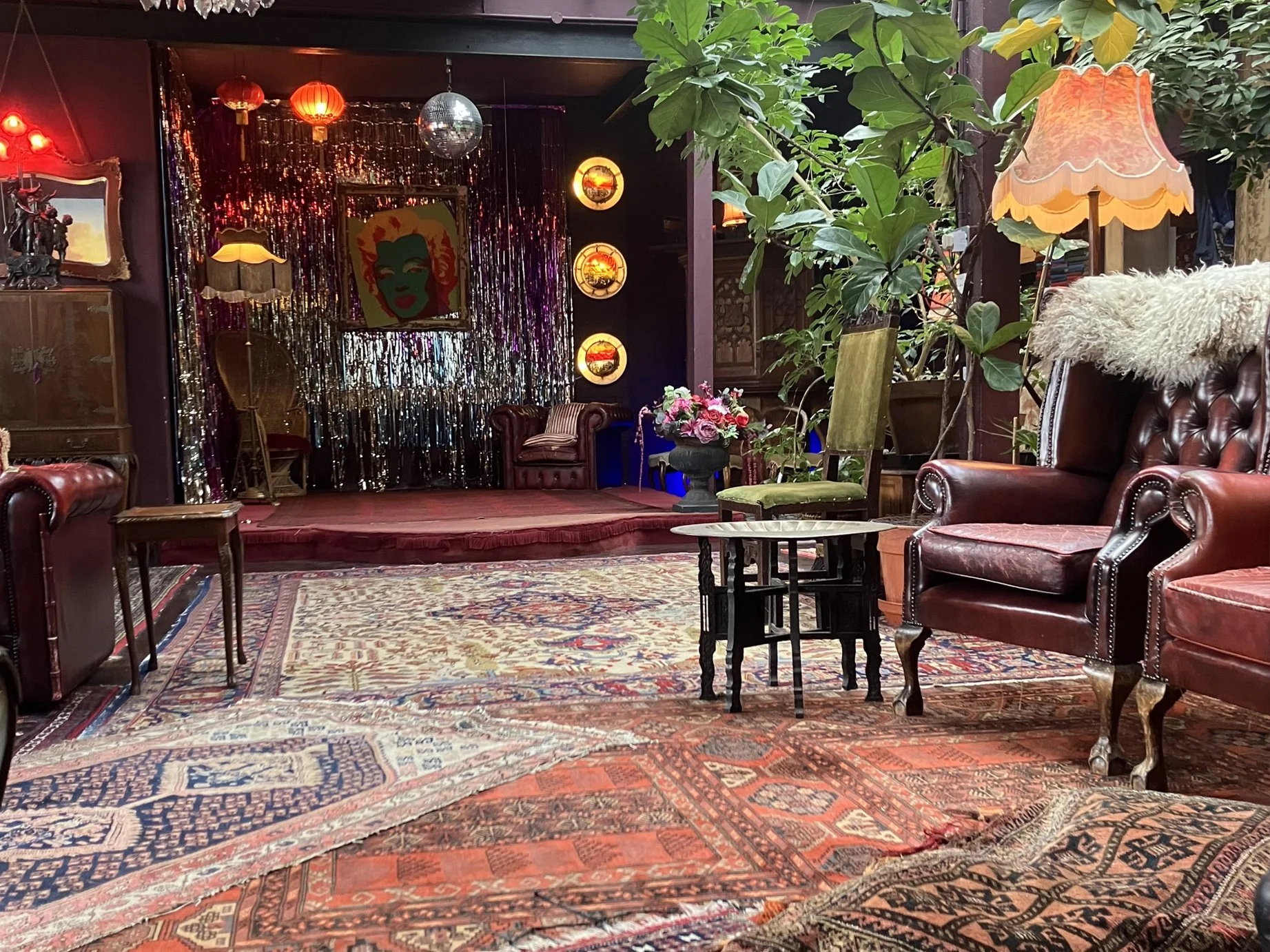 A cozy vintage-style lounge area with leather armchairs, a wooden side table, potted plants, and a small stage decorated with colorful lights, artwork, and metallic streamers. There is a lamp with a fringed lampshade and a floral arrangement on the s