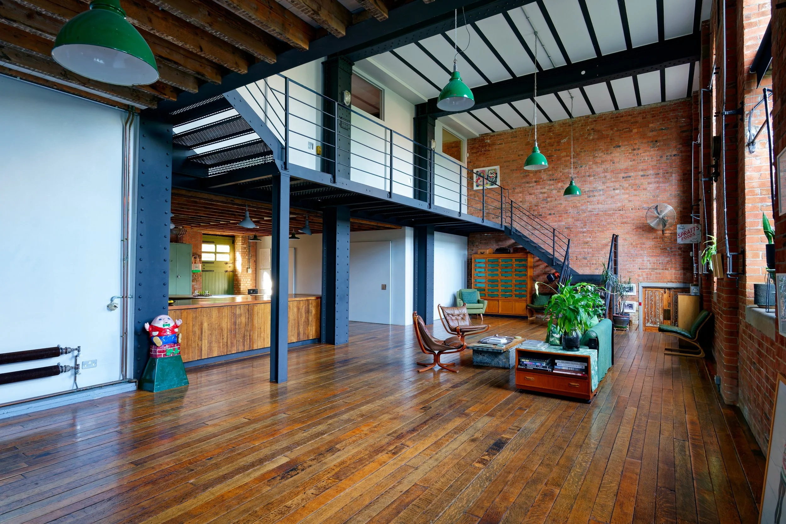 Industrial-style loft interior with exposed brick walls, wooden floors, a mezzanine with metal railing, hanging green pendant lights, and vintage furniture.