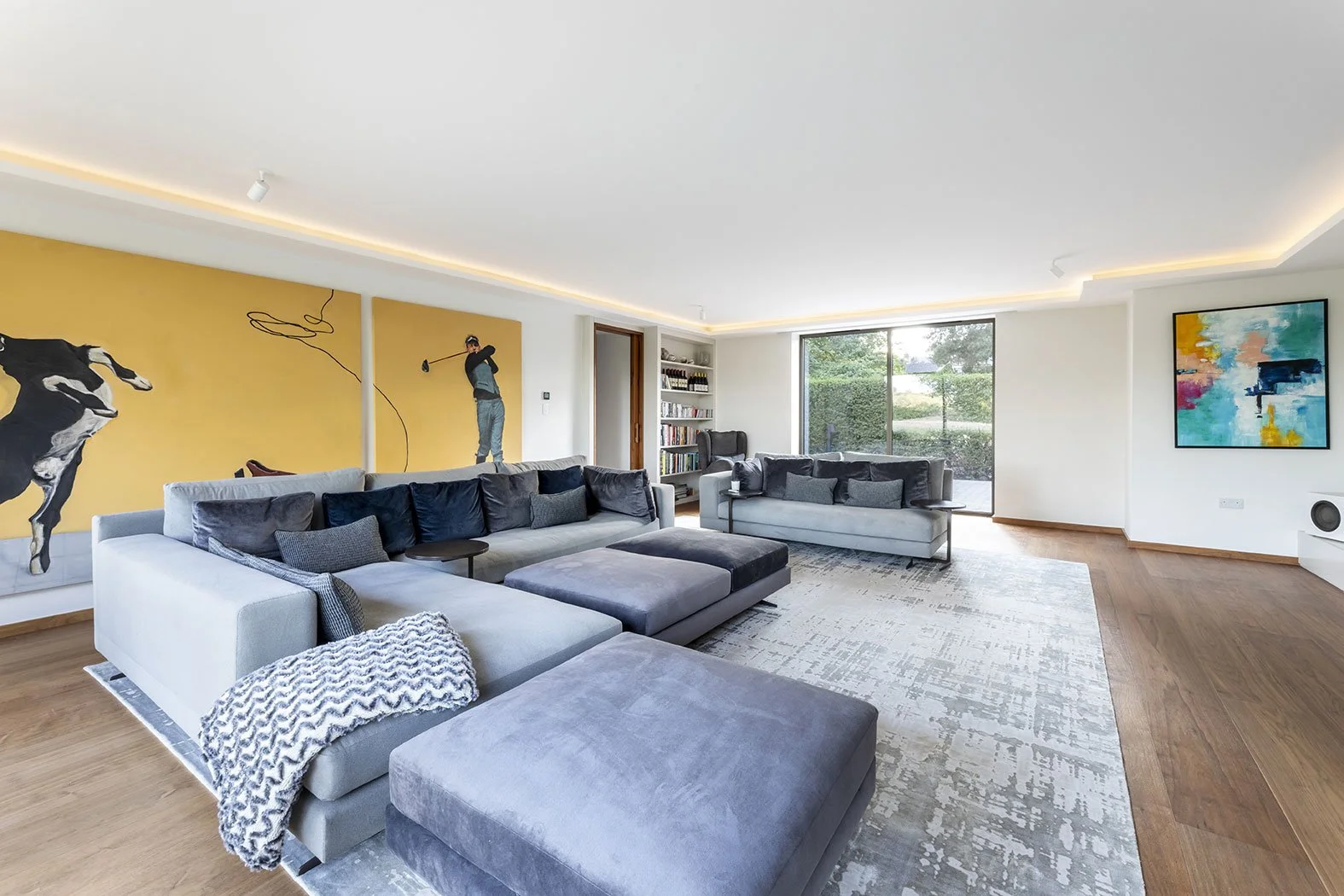 Modern living room with gray and black sectional sofas, abstract art on the walls, a large window overlooking greenery, and hardwood floors.