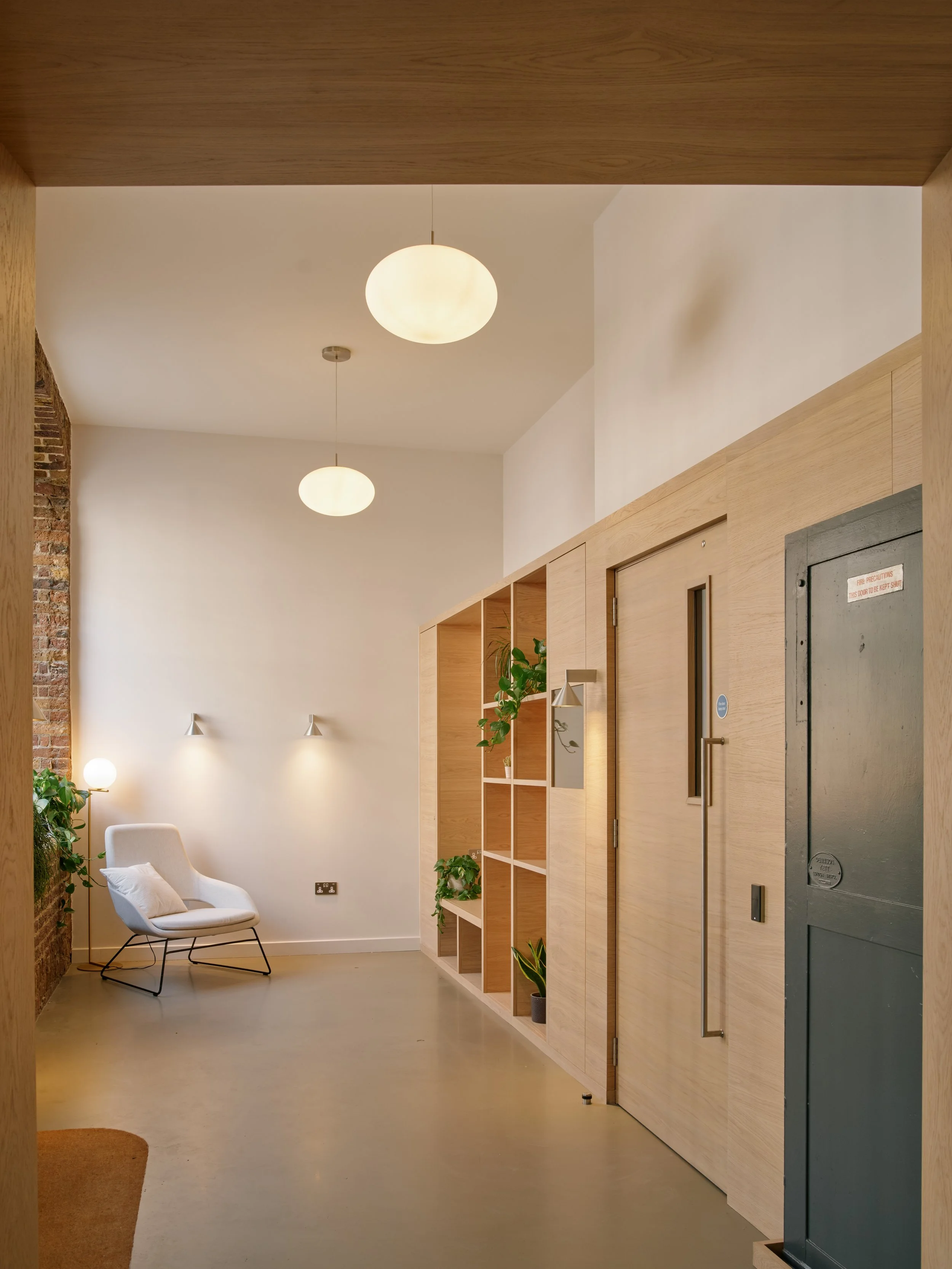 Modern interior space with a white accent wall, open wooden shelving with potted plants, a white armchair, wall-mounted lights, hanging globe ceiling lights, and a brick wall on the left.