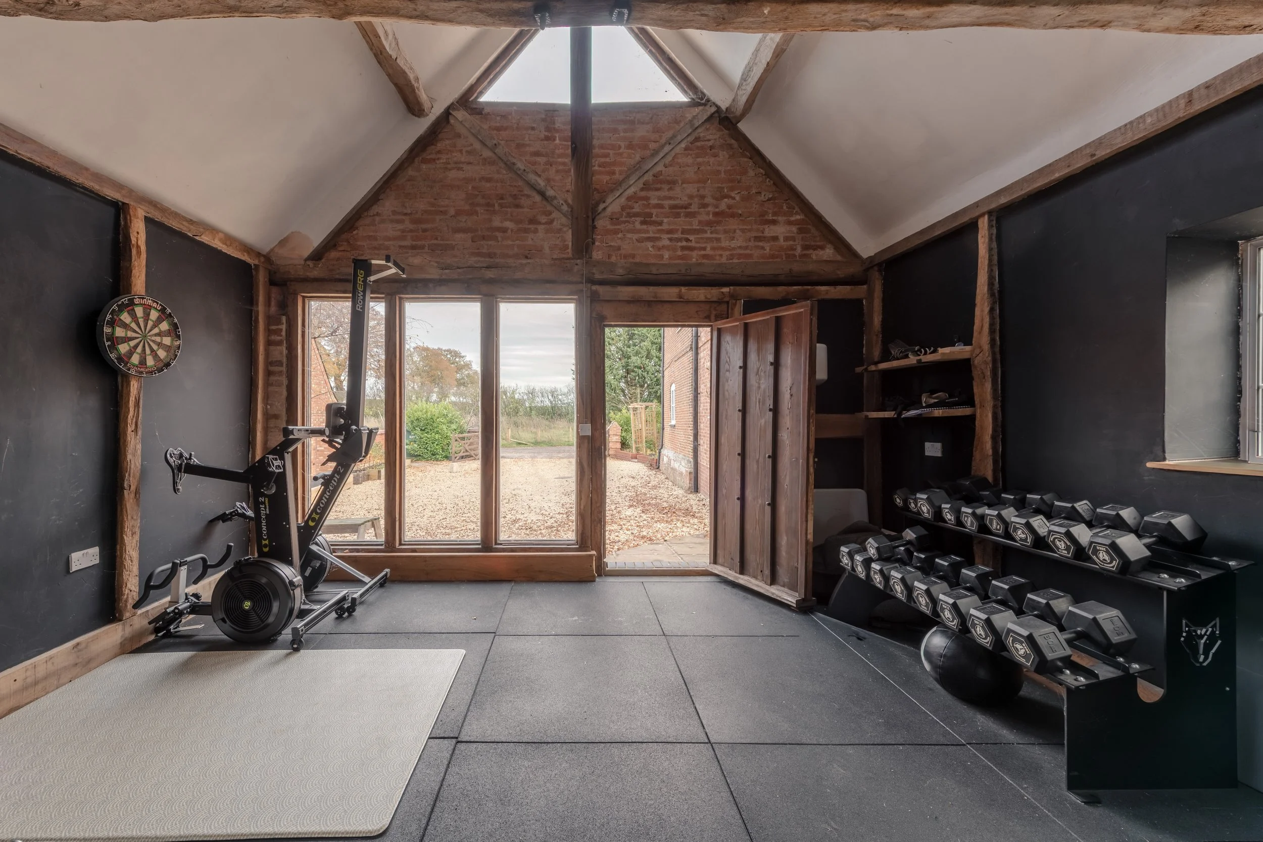 Home gym with black walls, wooden beams, window door leading outside, weight rack with dumbbells, exercise bike, and dartboard.