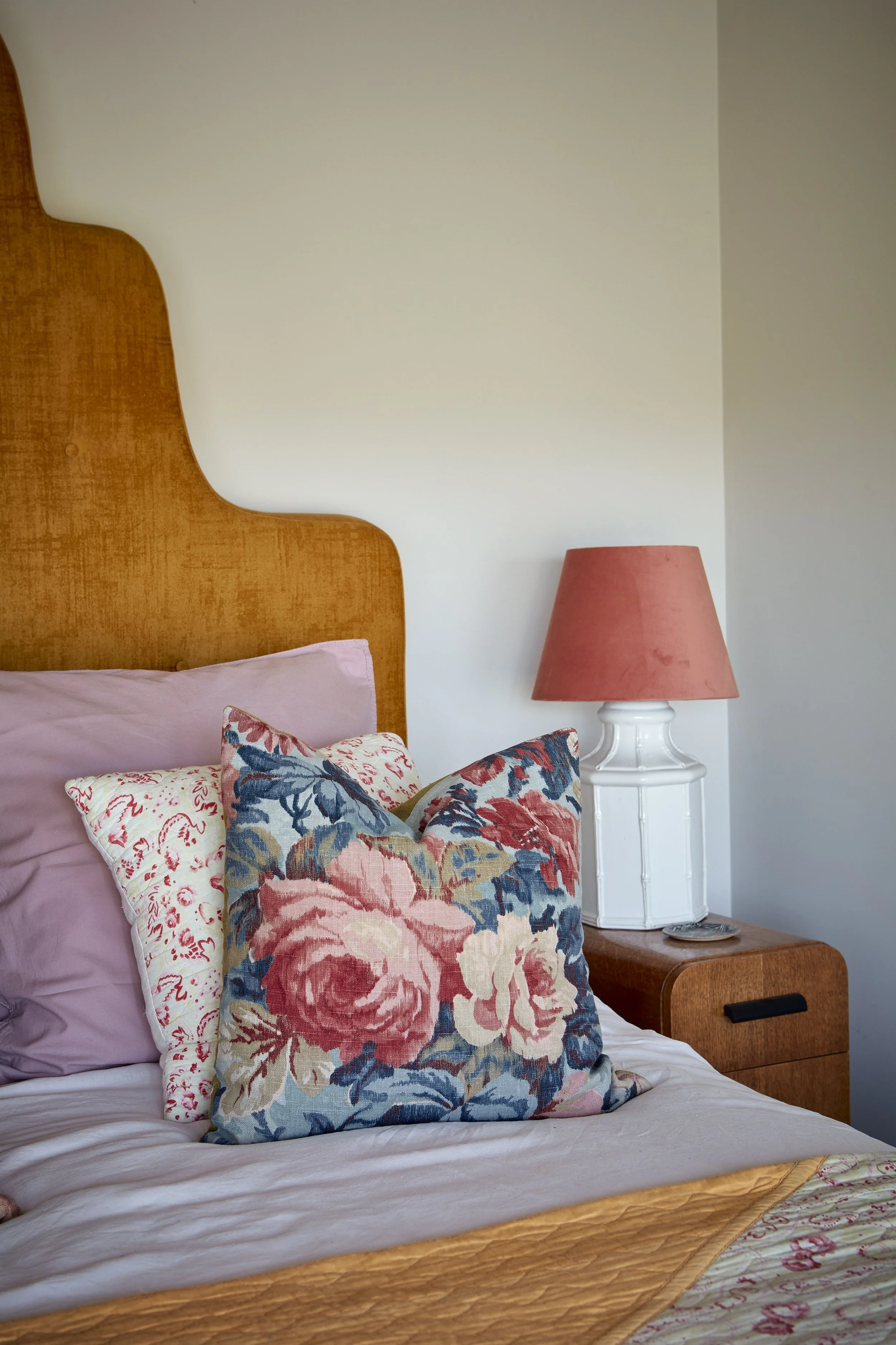 A bedroom with a wooden headboard, pink bed pillows, floral patterned throw pillows, a pink lampshade on a white bedside table, and a quilt with pink floral embroidery.