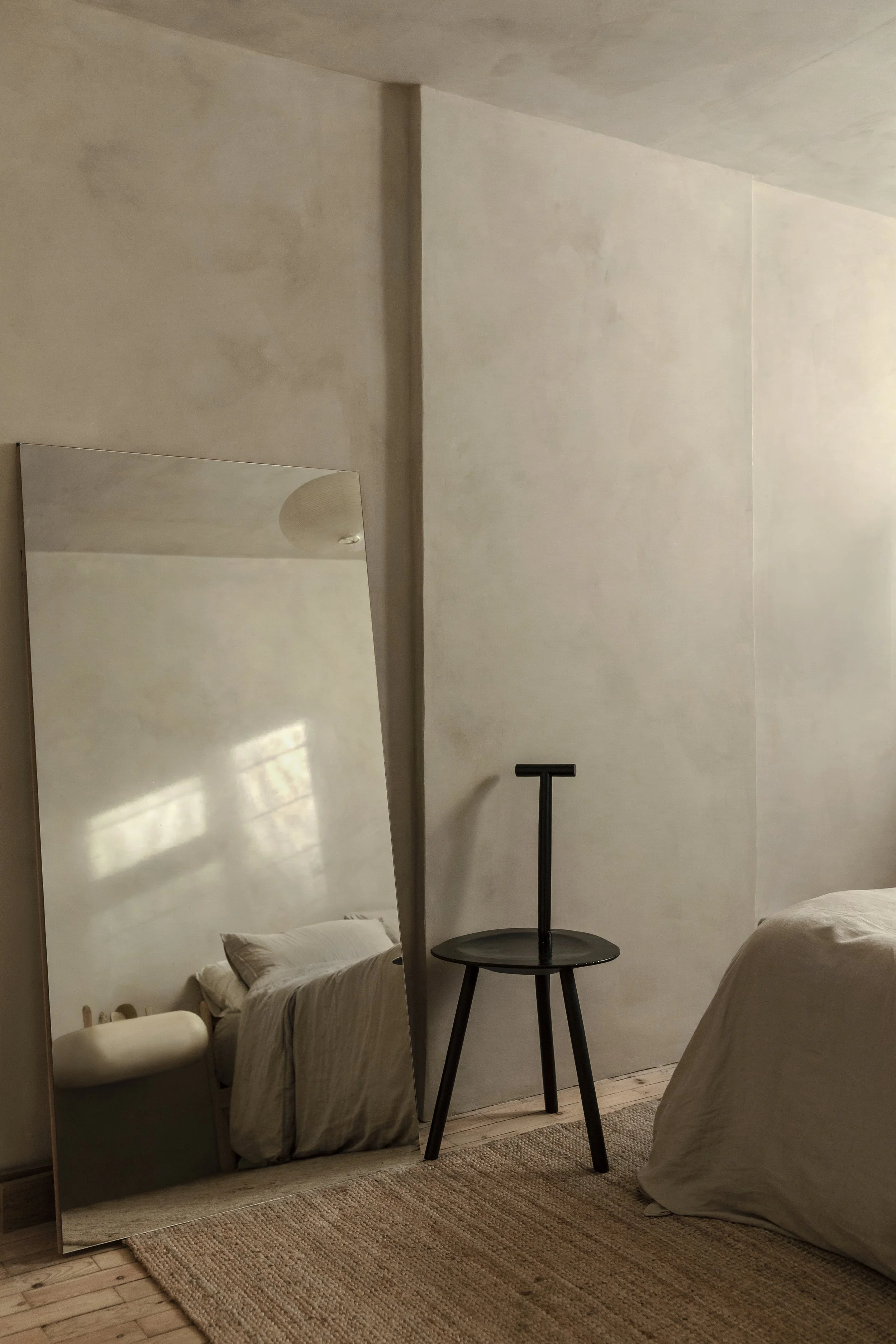 A minimalist bedroom corner showing a standing mirror reflecting a bed, a small table with a T-shaped handle on top, and part of the bed with a beige cover, against a plain off-white wall with a wooden floor and a woven rug.