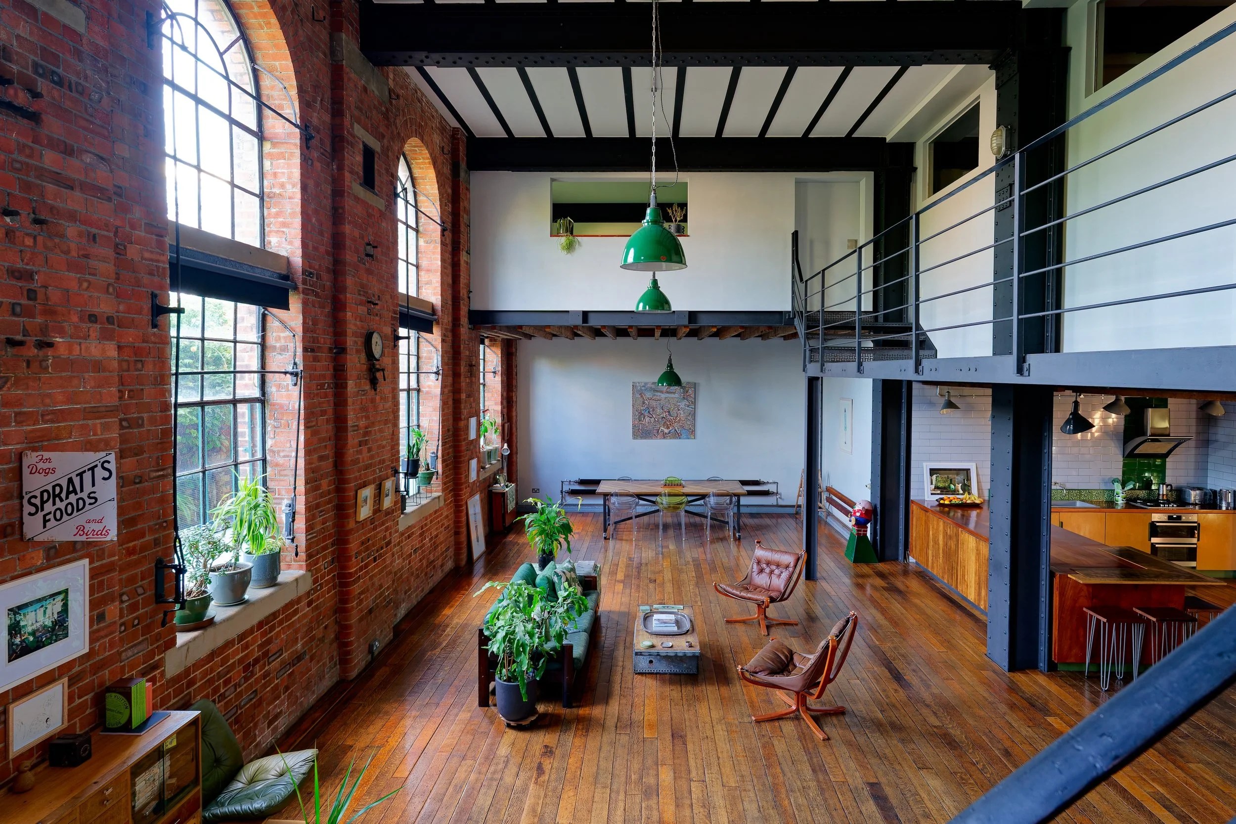 Spacious loft interior with red brick walls, large multi-pane windows, hardwood floors, and an open second level. Decor includes green pendant lights, potted plants, artwork, and mid-century modern chairs around a coffee table. Kitchen visible in the