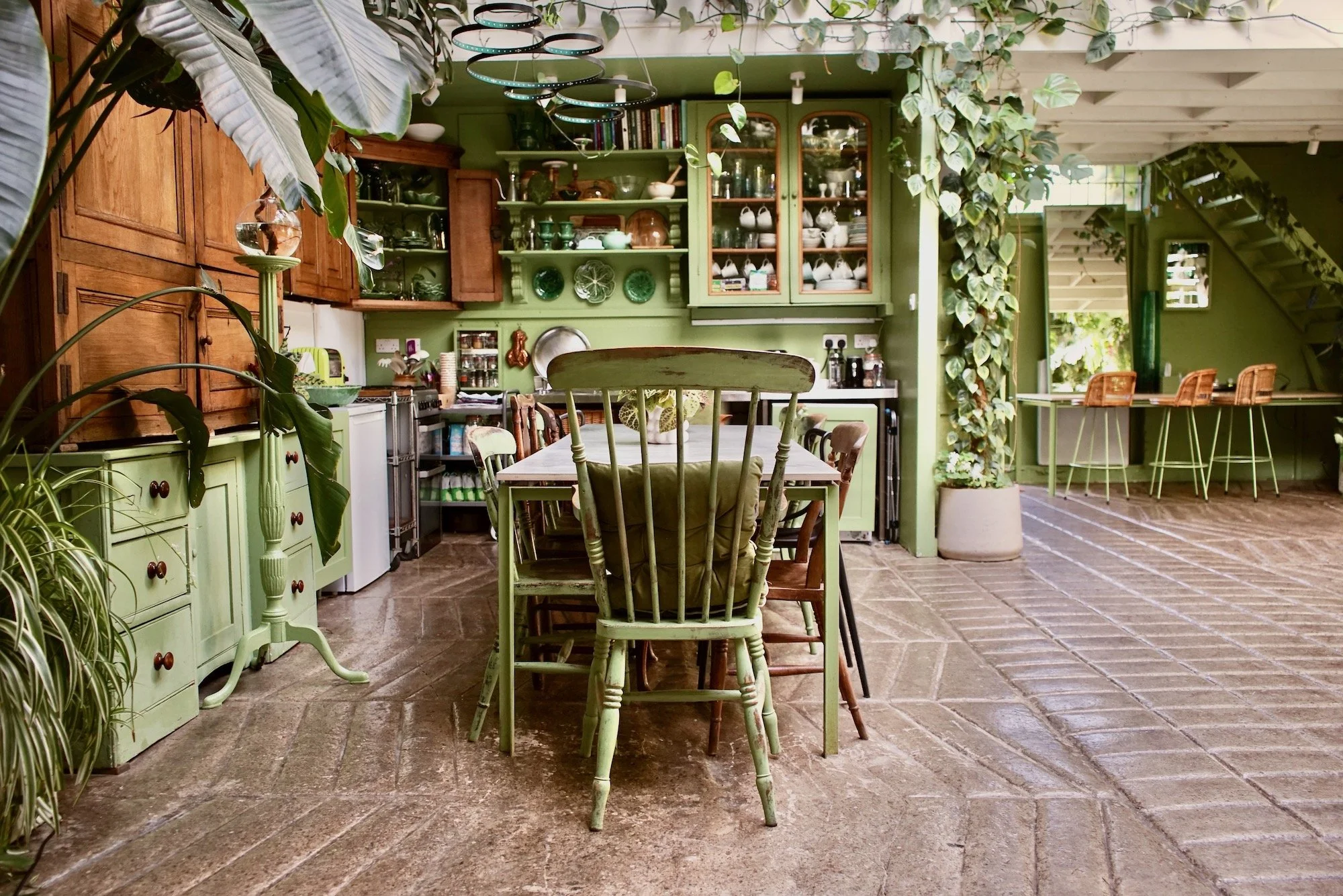 A cozy and colorful kitchen and dining area with green-painted walls and wooden cabinets, decorated with plants and various kitchenware, with a tiled floor and a central table surrounded by mismatched chairs.