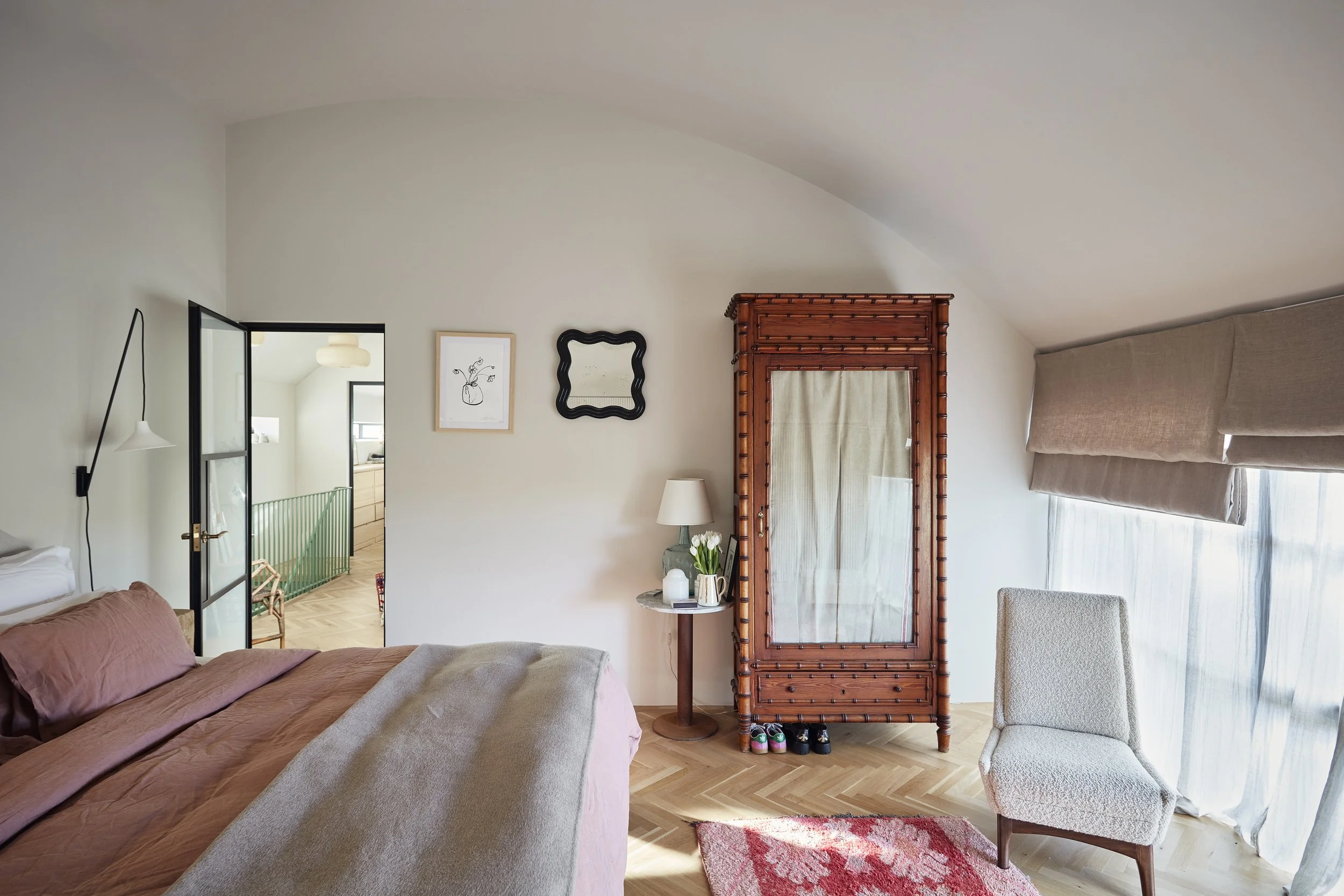 A cozy bedroom with a bed covered in pinkish-brown bedding, a small round side table with a lamp and flowers, a mirror, a framed artwork of a vase with flowers, a wooden wardrobe with a curtain, a light-colored armchair, and a window with beige curta