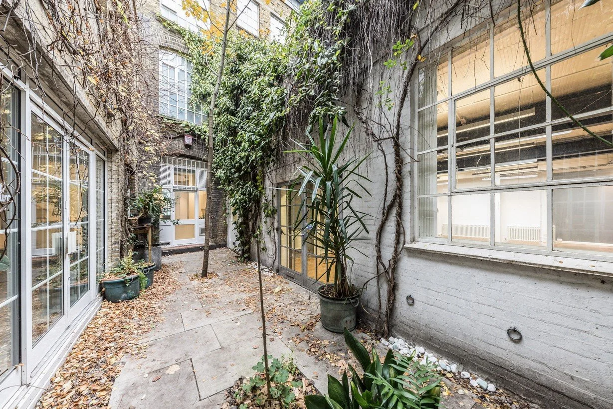 A small outdoor courtyard or patio with potted plants, leaf-covered stone tiles, surrounded by brick walls with large windows, some vines growing on the walls, and an autumnal atmosphere.