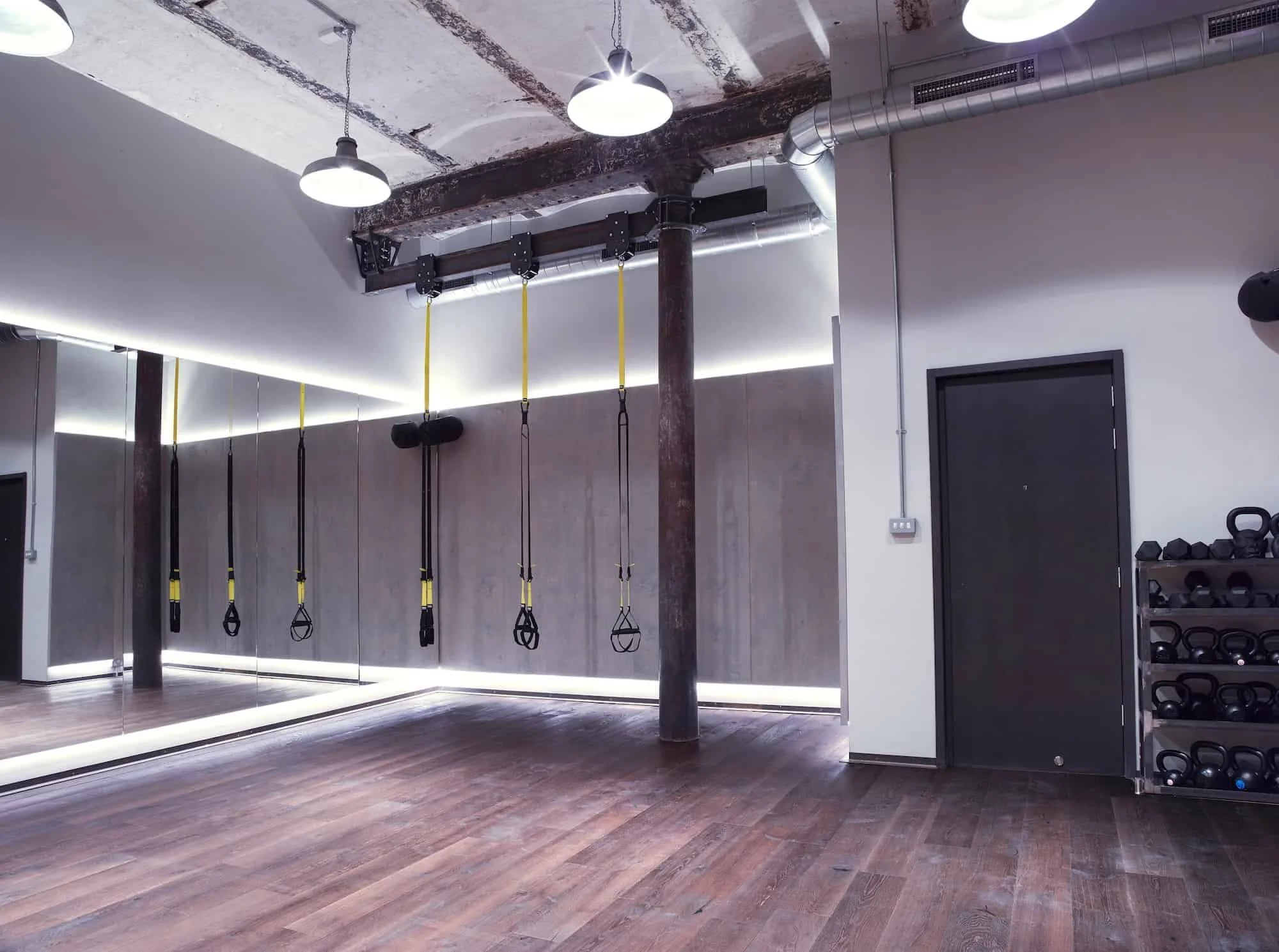 Empty modern fitness studio with wooden floors, wall mirrors, hanging TRX suspension straps, and dumbbell racks.