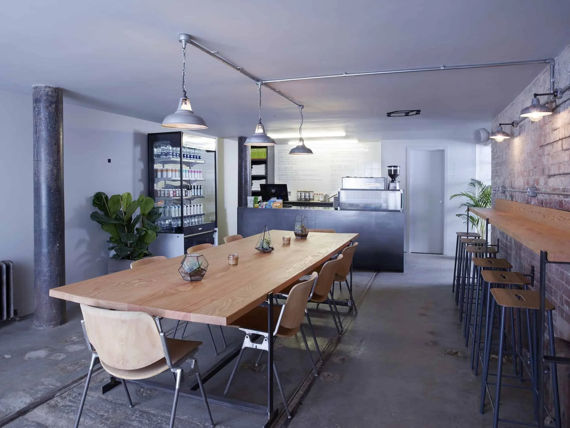 Interior of a modern cafe or coffee shop with a long wooden table surrounded by chairs, a barista counter in the background, a refrigerator with drinks, and decorative plants on the side. The space features an industrial style with concrete floors, e