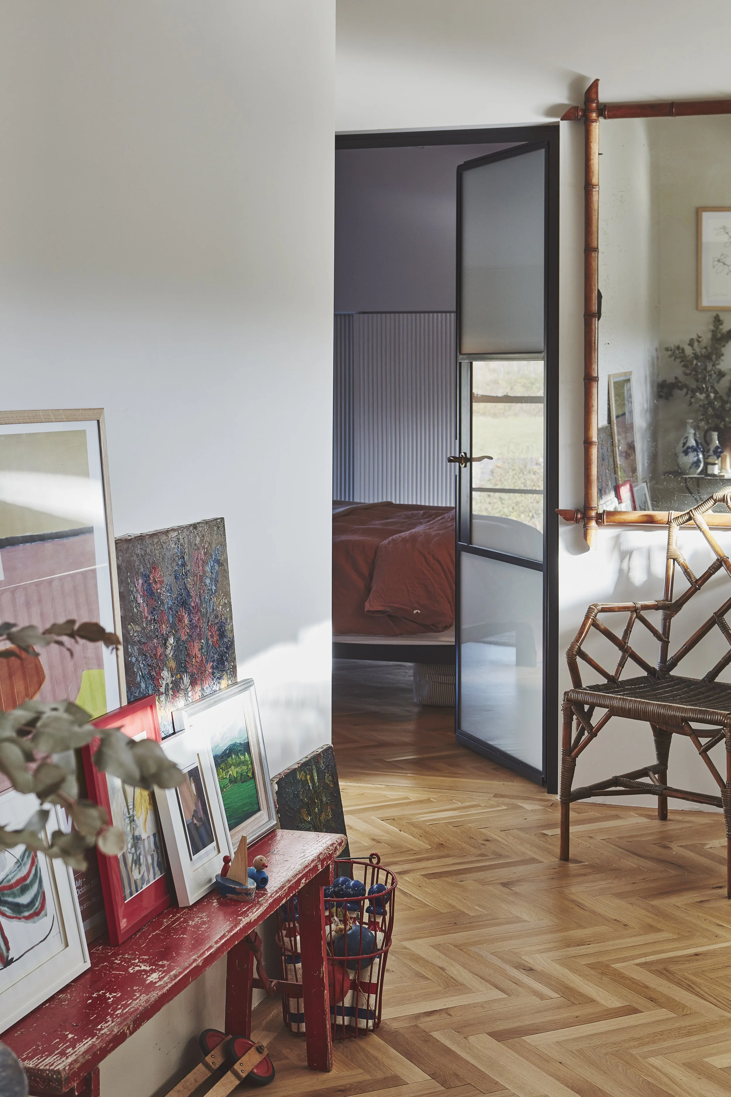 Interior of a room with a hardwood floor, artwork displayed on a red painted console table, and a glass door leading to a bedroom. The bedroom has an orange bedspread, and natural light is coming through the window. There is a wooden and wicker chair