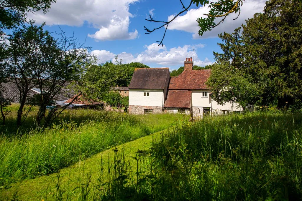 A country house with white walls and red roof tiles, surrounded by tall green grass and trees, under a partly cloudy blue sky.