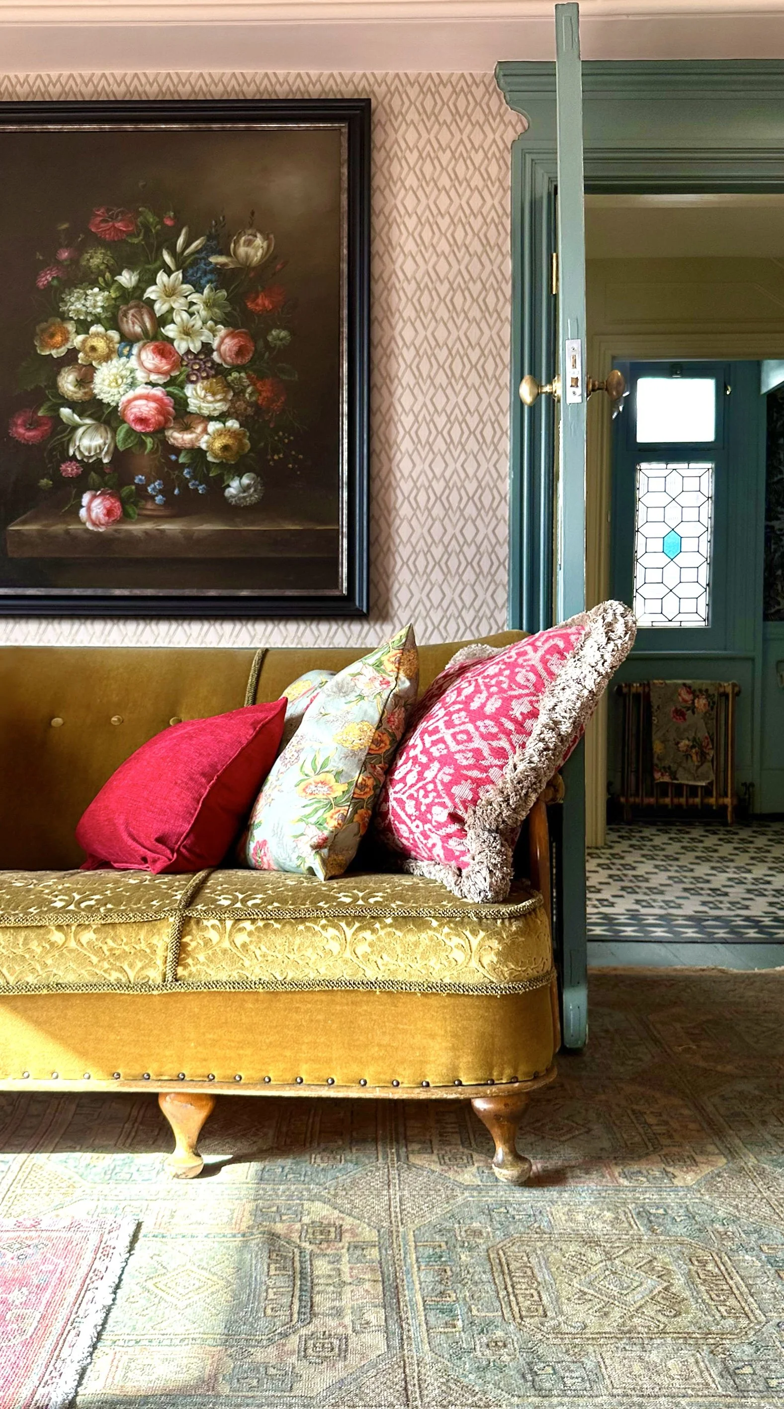 A vintage yellow velvet sofa with colorful pillows, positioned in front of a floral still life painting on the wall, with an open doorway leading to a hallway with patterned flooring.