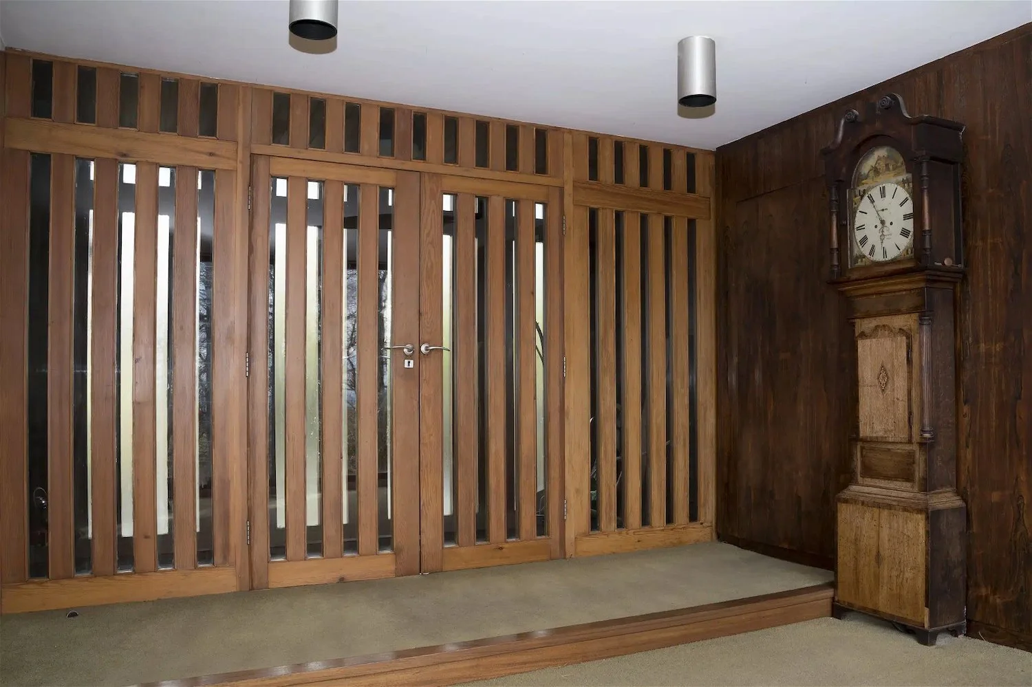 Interior of a room with wooden double doors and wooden wall panels, a vintage grandfather clock, and two ceiling lights.