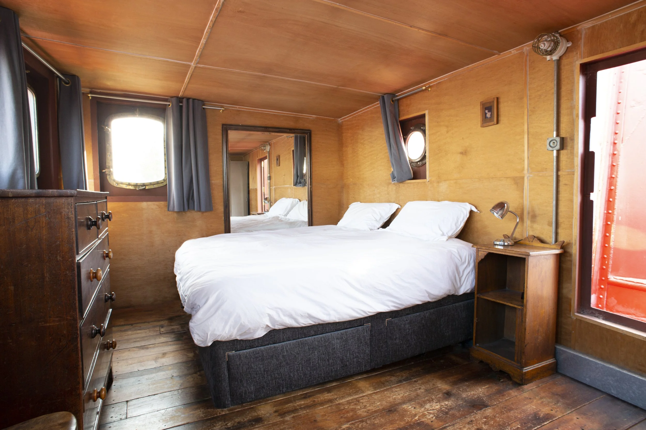 A cozy bedroom with wooden walls and ceiling, featuring a large bed with white bedding, a bedside table with a small lamp, and a tall wooden dresser. There are small windows with dark curtains and a mirror on the wall reflecting the room.