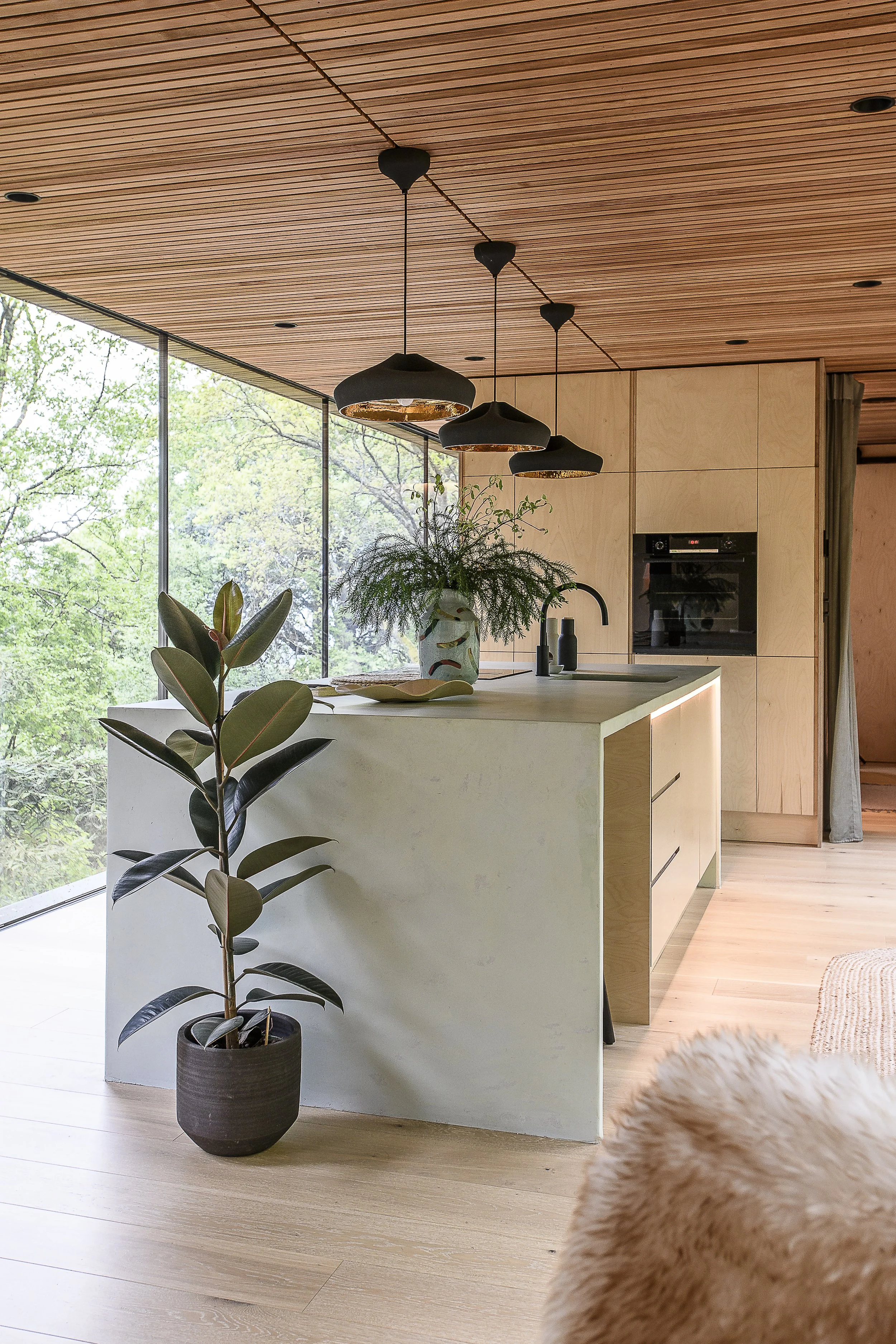 Interior view of a modern kitchen with a white island, black hanging pendant lights, a built-in oven, and a large window showing green trees outside. There are decorative plants inside the kitchen.