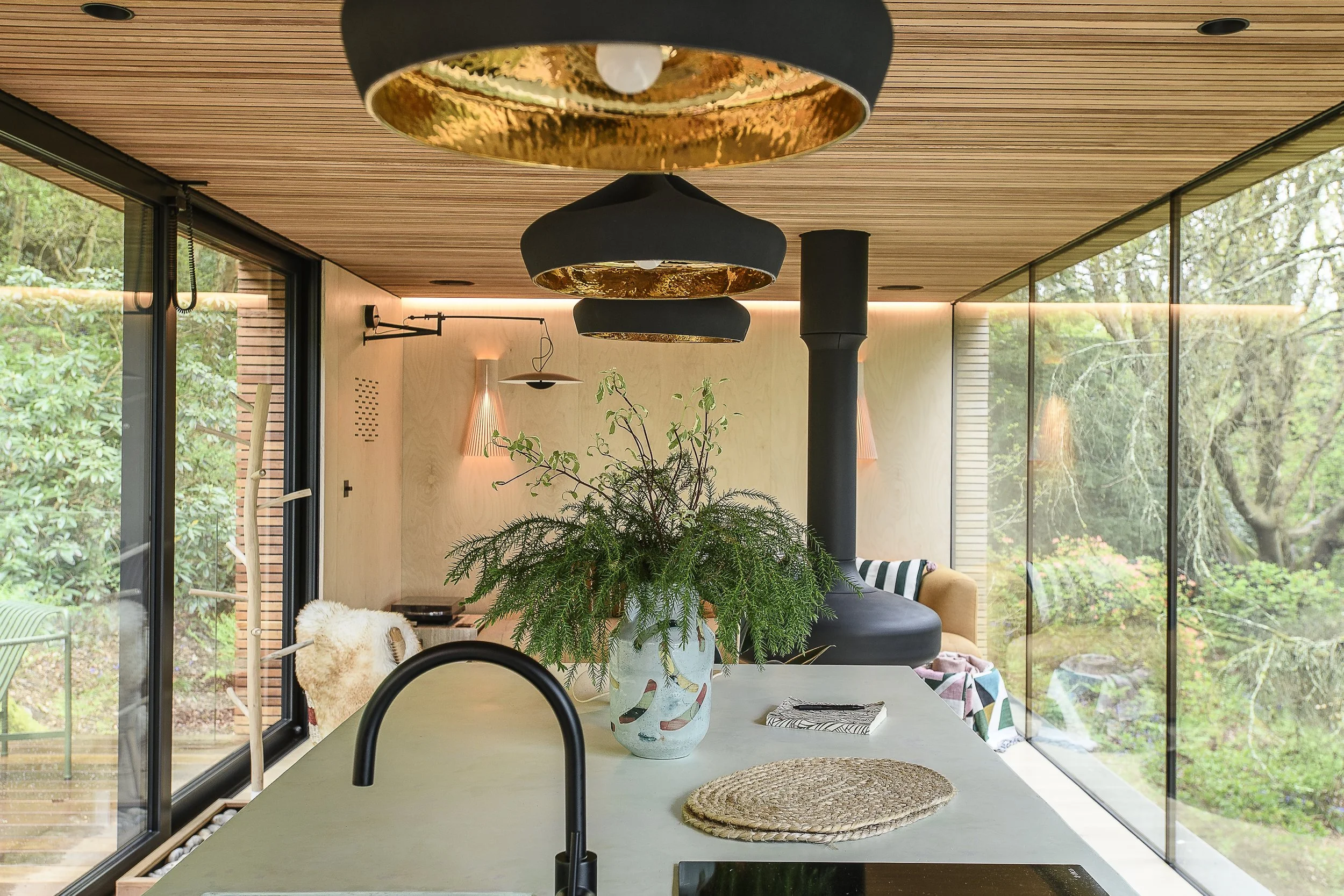 Modern kitchen with large glass windows showing greenery outside, a white countertop with a potted plant in a colorful vase, a woven mat, a notebook, and a stove; space features black and gold pendant lights and wooden ceiling.