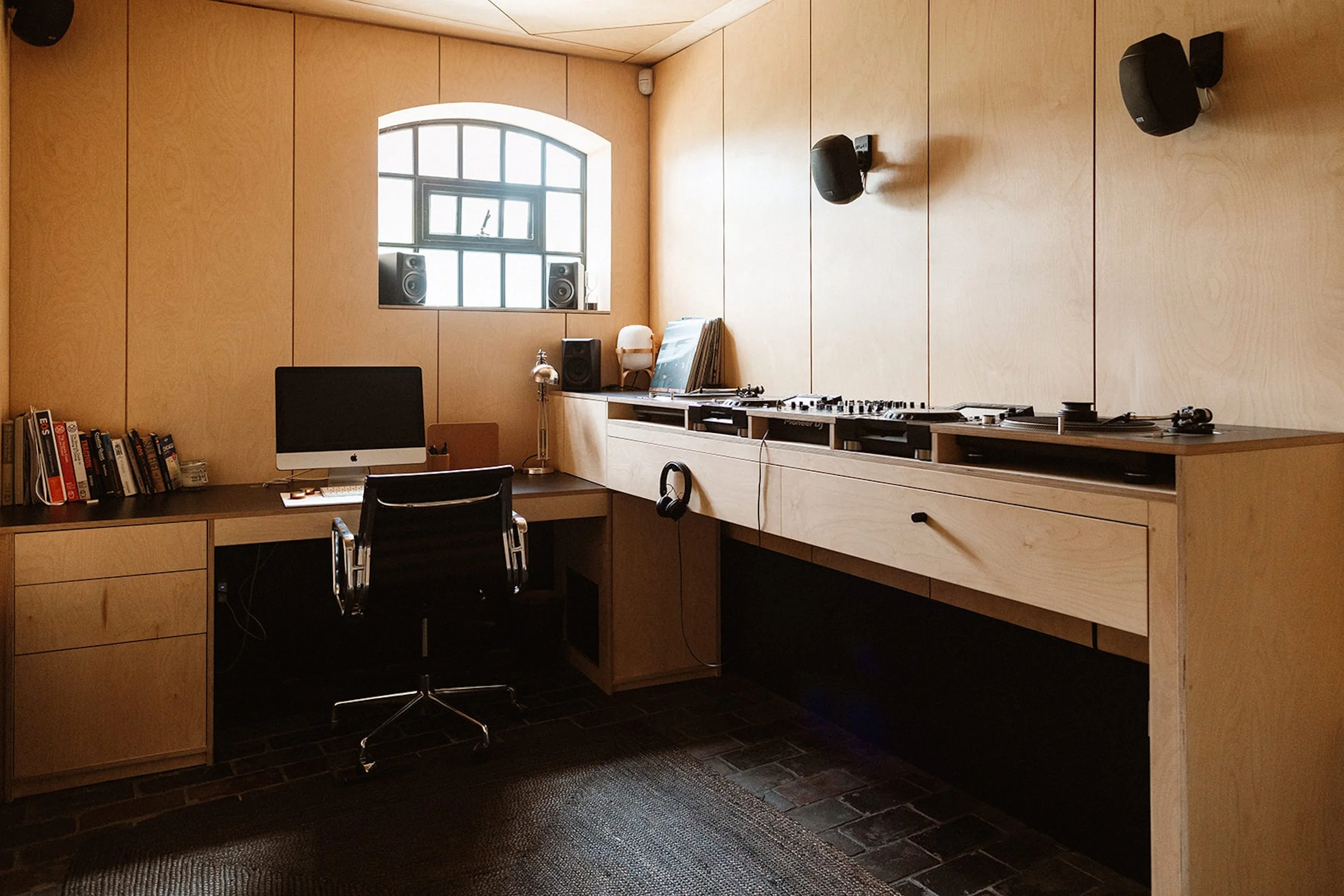 A minimalistic music production studio with a wooden desk holding an iMac computer, books, and a lamp, with a DJ controller on the right side, a black office chair, wooden wall panels, a small window with black speakers, and hanging black speakers on