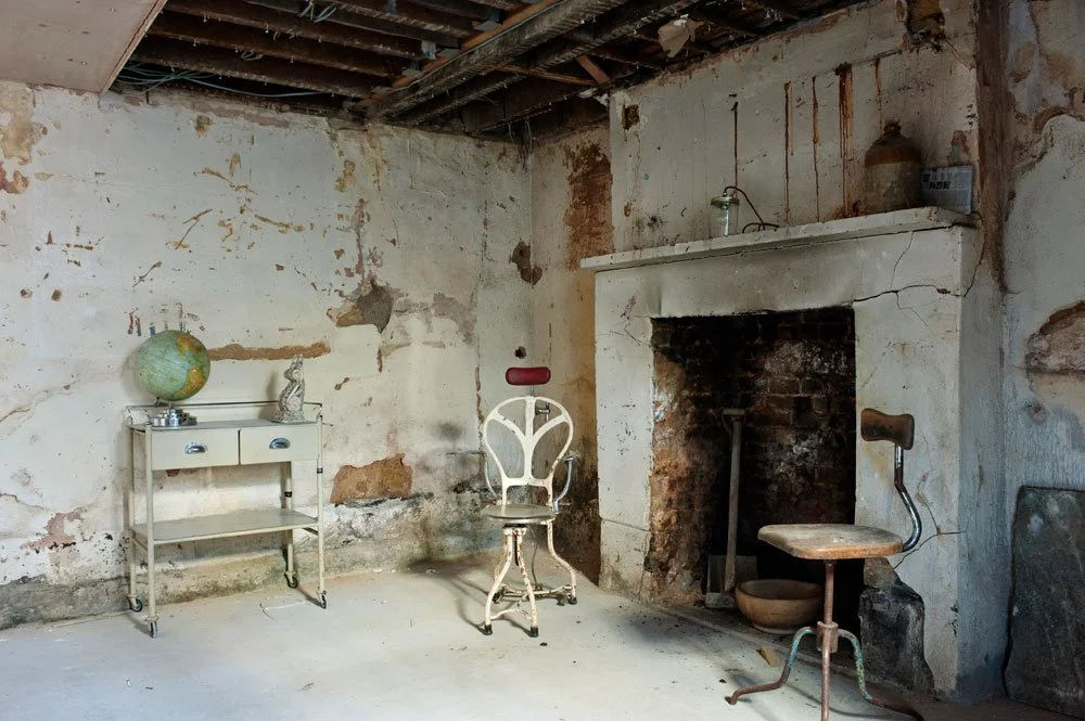 Old, dilapidated room with worn walls, exposed ceiling beams, and vintage furniture including a globe, a small table, and two old chairs.