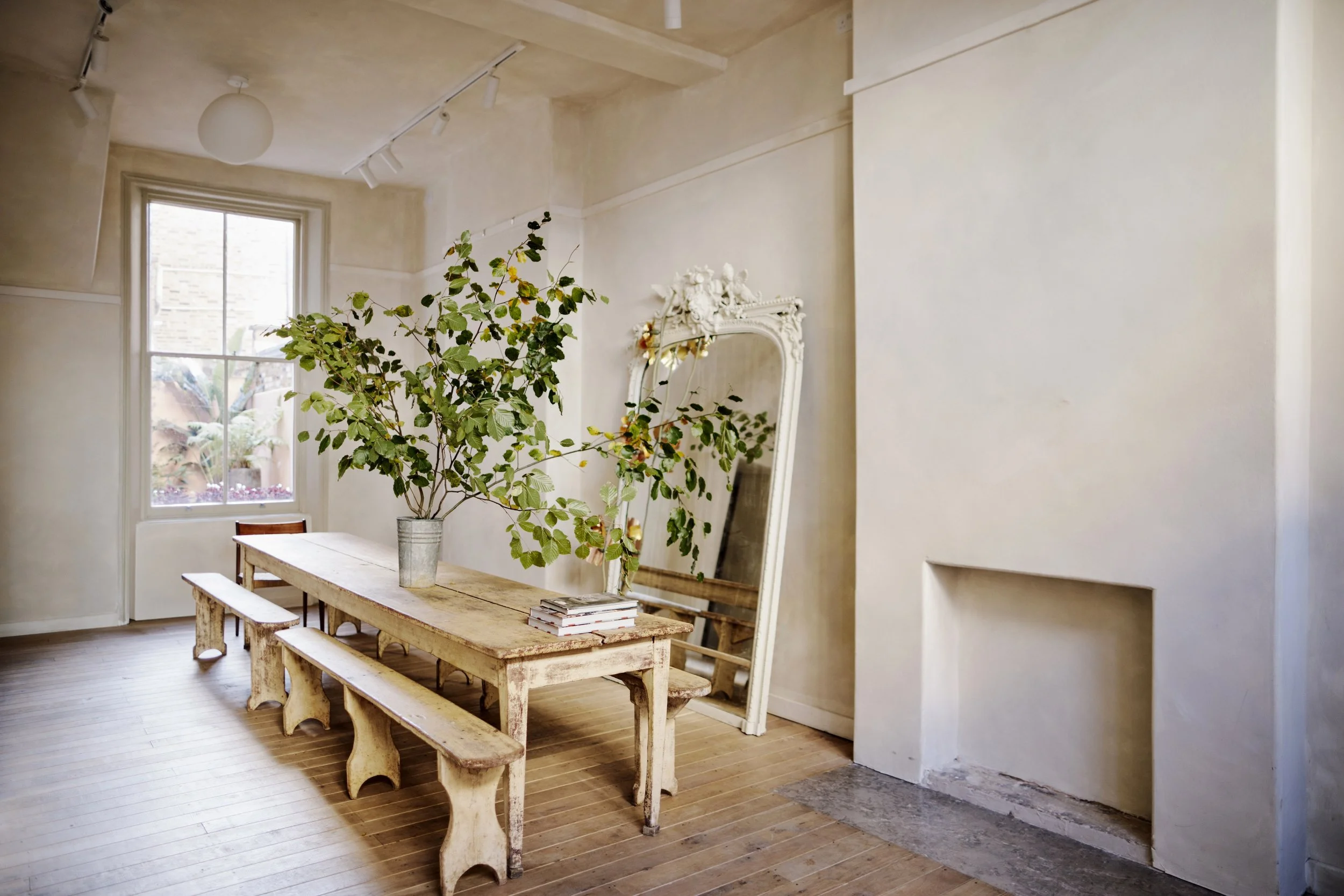 A minimalist interior with a wooden dining table and two matching benches, a large green plant in a metallic vase, a vintage ornate white mirror leaning against the wall, a window with a view of outdoor greenery, and a fireplace opening with exposed 