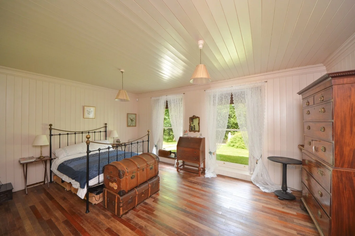 A cozy bedroom with wooden floors, two large windows with lace curtains, a metal bed with white bedding and navy blue blanket, vintage chest at the foot, wooden dresser, small writing desk, and corner table. Green trees visible outside.