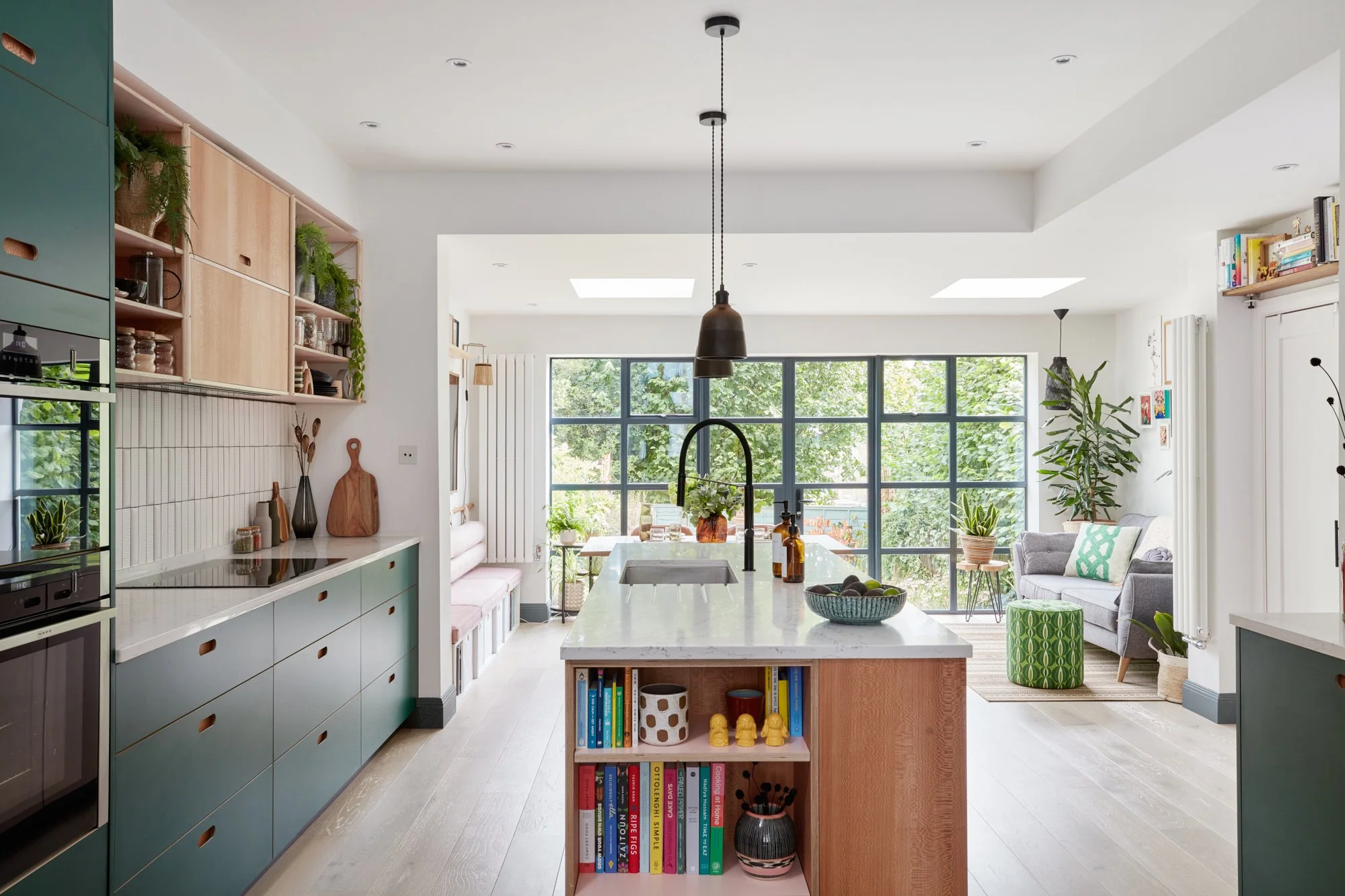 Bright, modern kitchen with an island in the center, featuring a sink and decorative jars, surrounded by green cabinets, open shelving with plants, and a sitting area with a sofa and large windows overlooking greenery.