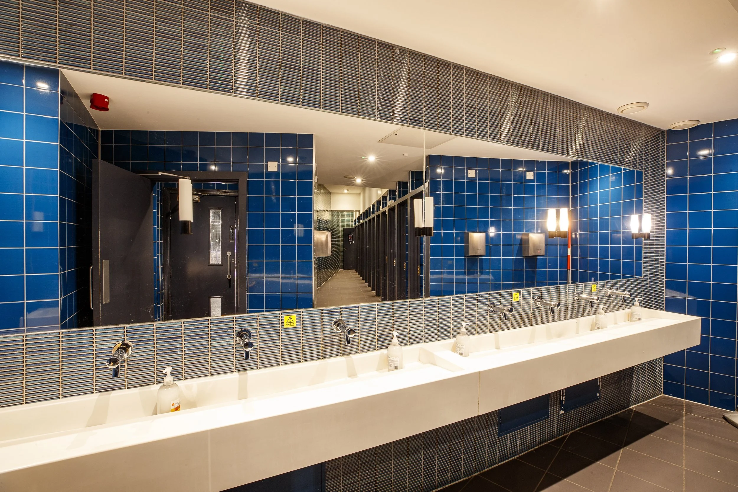 Public restroom with a long white sink, six faucets, hand soap bottles, and a large mirror reflecting blue tiled walls and multiple bathroom stalls.