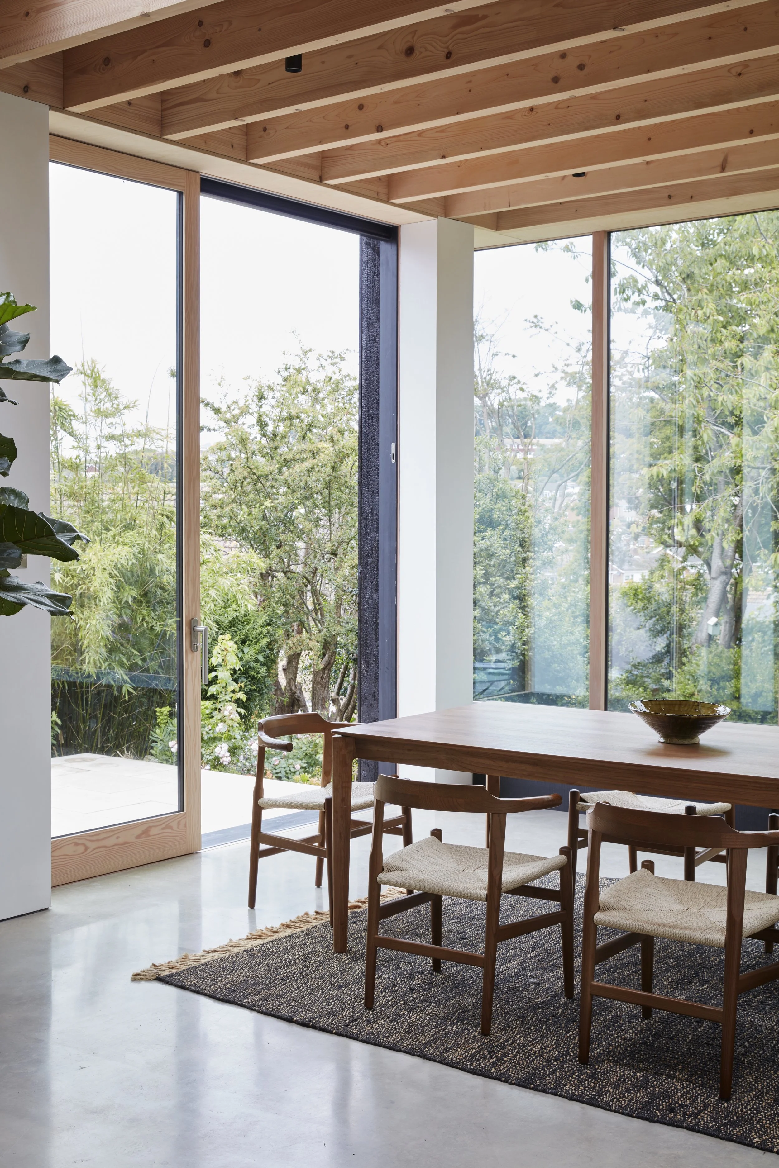 Bright dining area with a wooden table, four chairs with woven seats, a black rug, and large corner windows showing greenery outside.