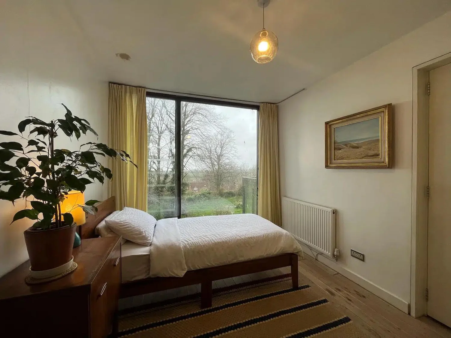 A bedroom with a large window showing an outdoor view with trees, a bed with white linens, a wooden side table with a potted plant, and a painting on the wall.