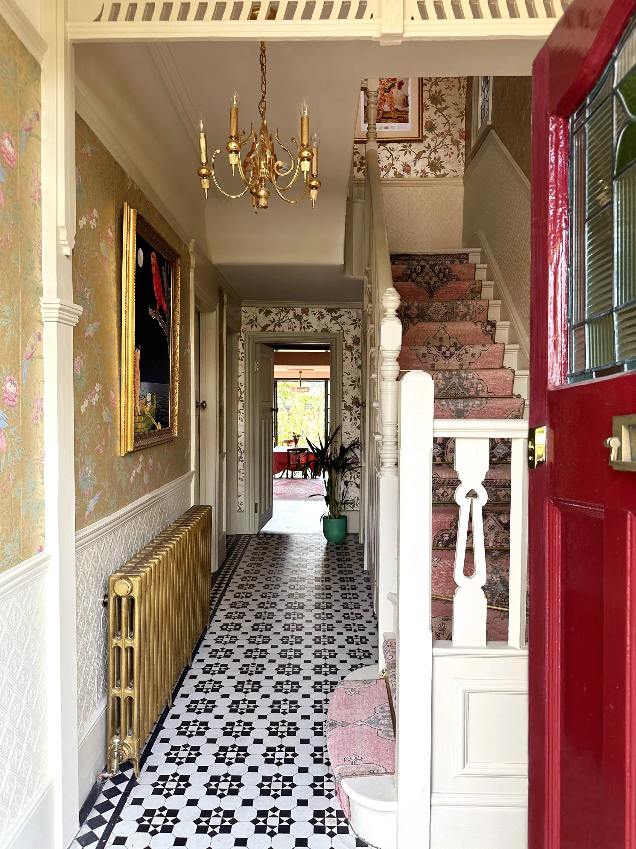 View of an entry hall in a house featuring a black and white patterned tile floor, a gold radiator, floral wallpaper, a chandelier, a stairway with a pink patterned runner, and a glimpse into a dining room with a red tablecloth.