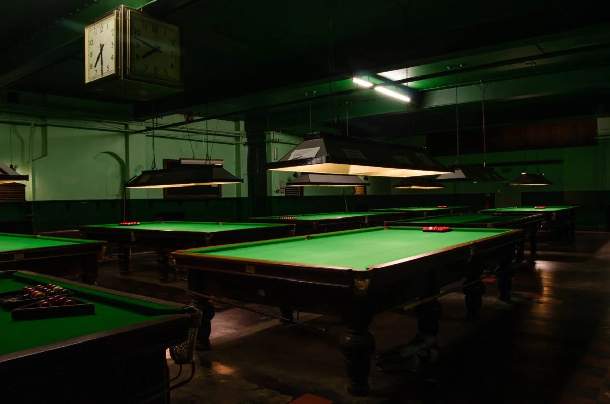 Multiple snooker tables with green felt surfaces and overhead lighting in a dimly lit pool hall