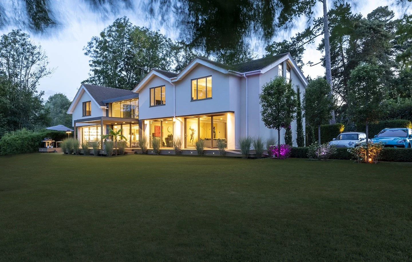 A modern two-story house illuminated at dusk, surrounded by a well-maintained lawn, trees, and parked cars, with warm interior lights visible through large windows.