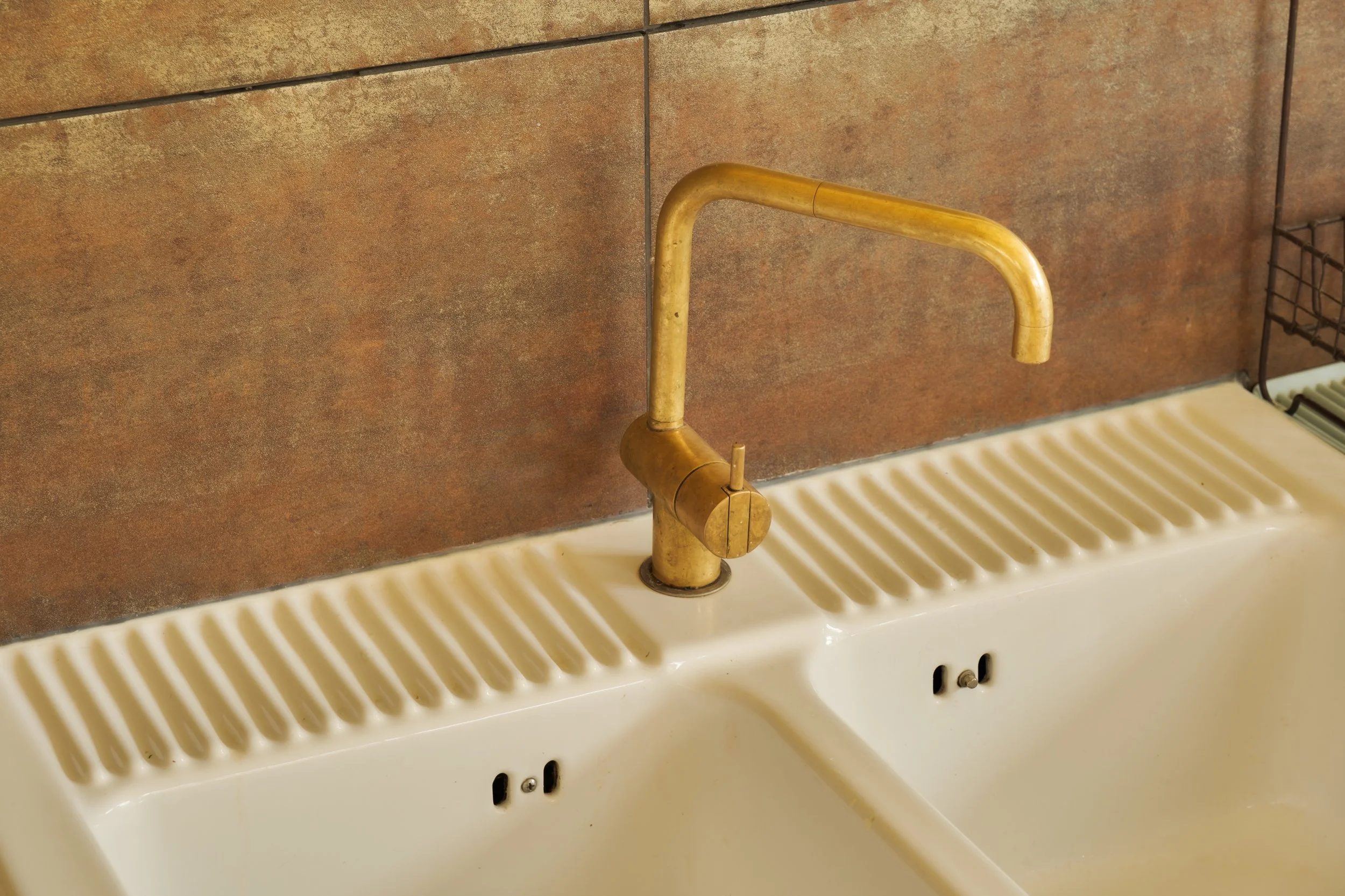A white ceramic kitchen sink with a brass faucet and a textured draining area, mounted against a brown tiled wall.