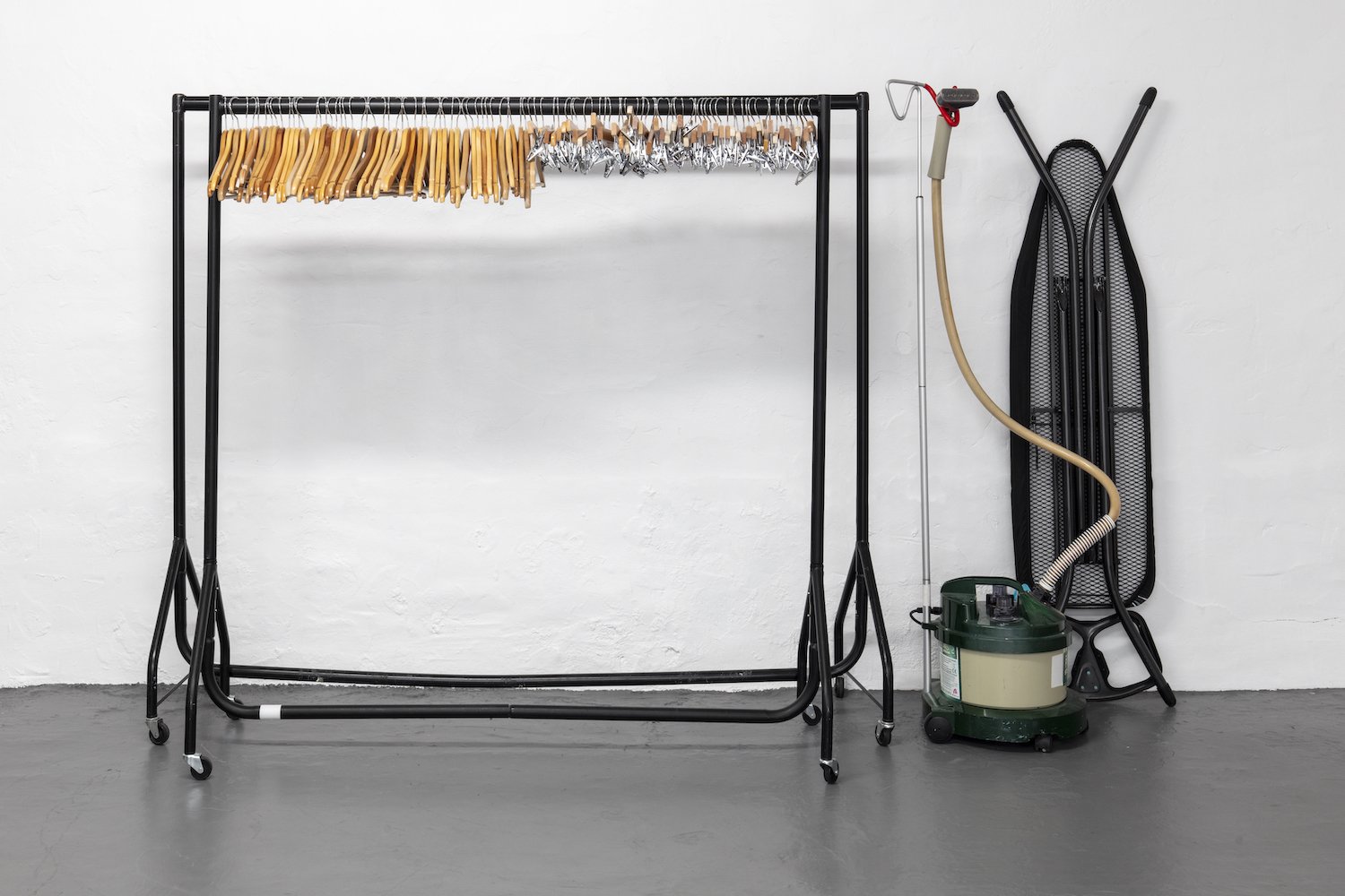 Laundry room with a metal clothing rack holding hangers, a handheld vacuum, a steam mop, and a black mesh storage case leaning against a white wall.