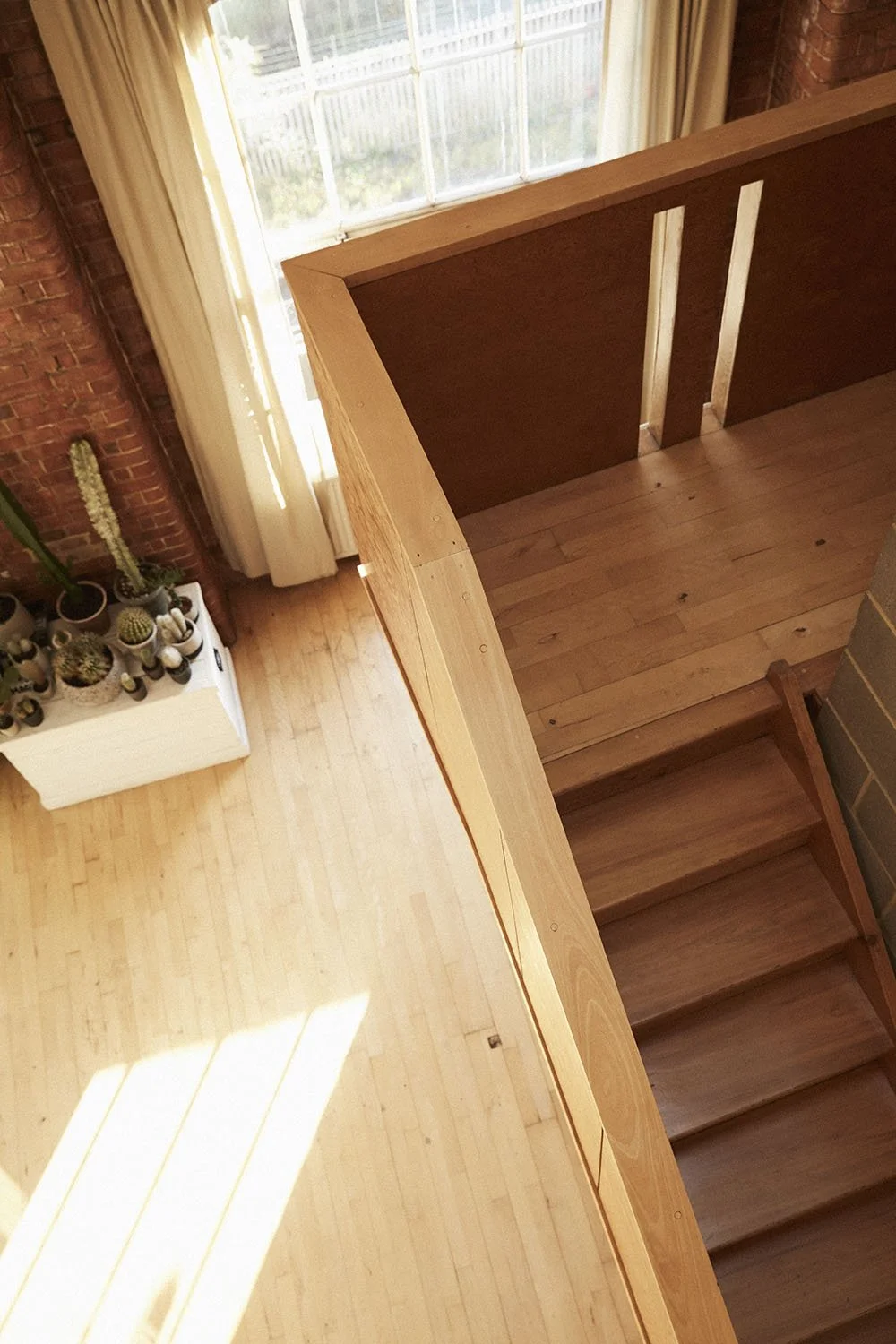 Interior view of a wooden staircase leading down to a sunlit room with a white cabinet holding potted cacti and succulents, beige curtains, brick wall, and a large window.