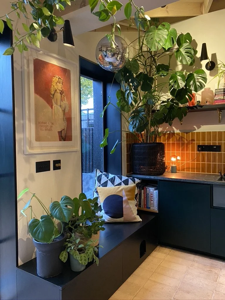 A cozy indoor space with a large leafy plant, a disco ball hanging from the ceiling, and a black bench with potted plants, a decorative cushion, and books. The wall has a framed artwork, black wall sconces, and a small orange and yellow tiled counter