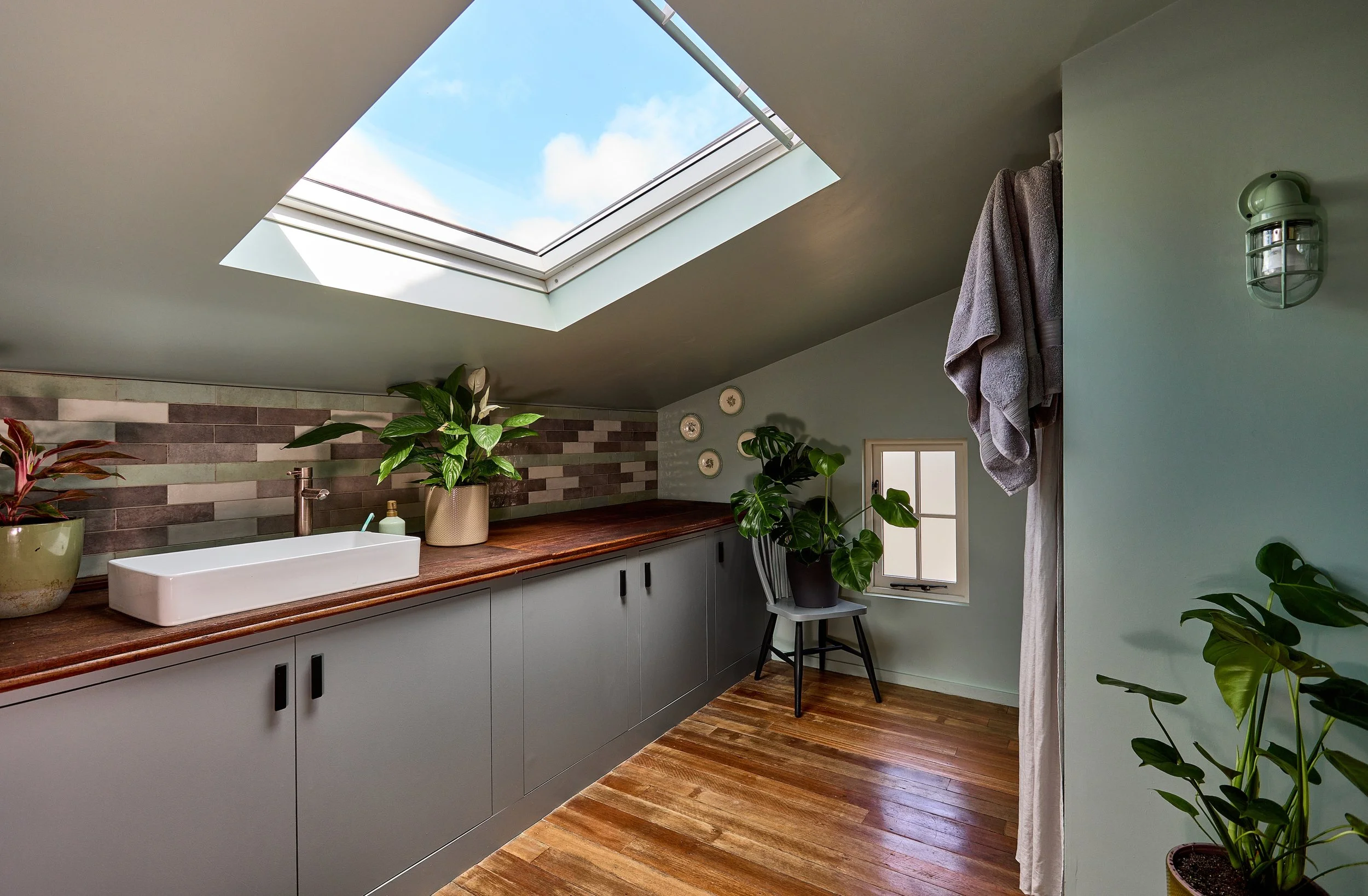 Attic bathroom with skylight, green walls, and wooden floor, featuring a sink with plants, a small window, and hanging towels.