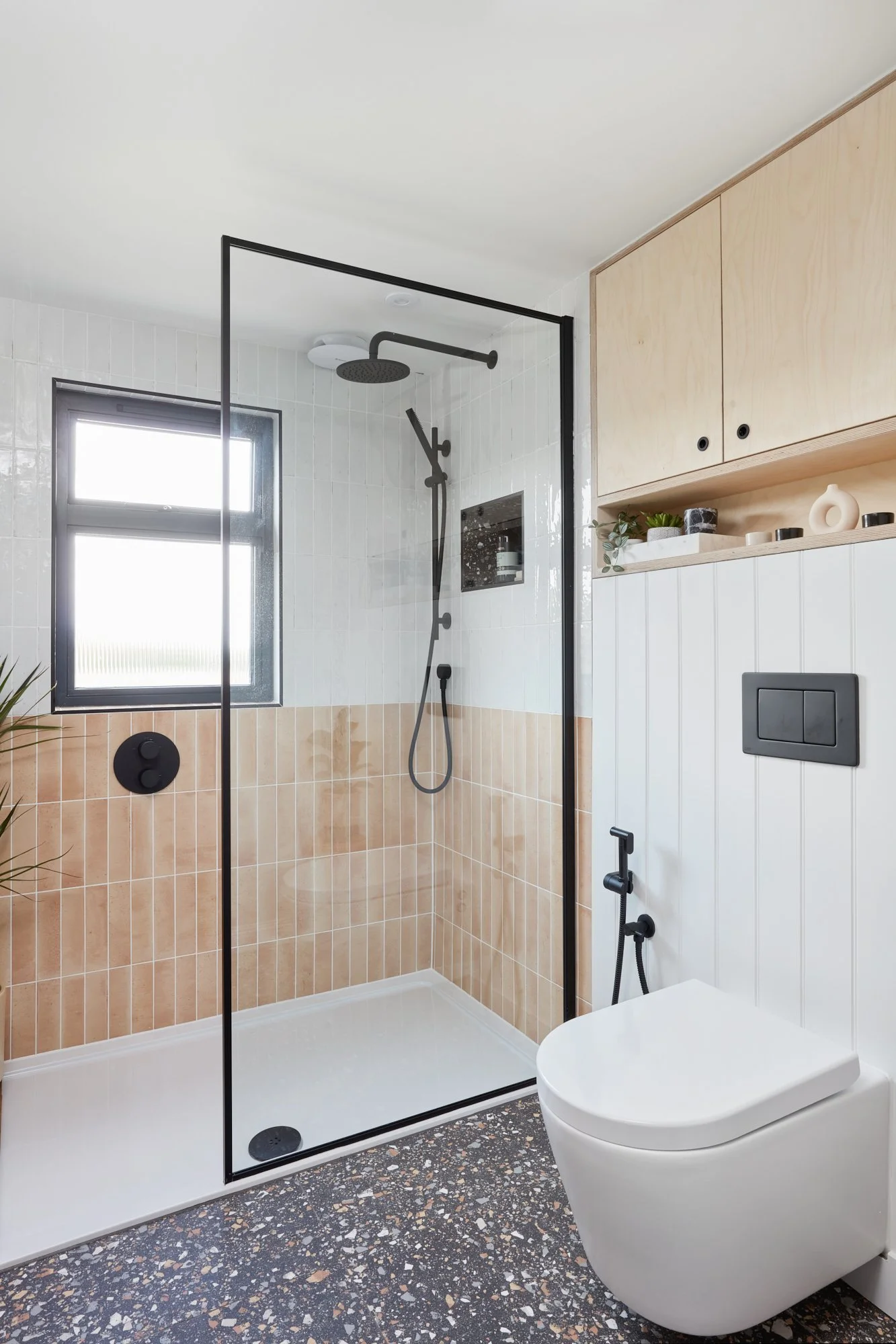 Modern bathroom with a walk-in shower with black framing, beige tiled lower walls, and white upper walls. Includes a black showerhead, window, ceiling light, toilet, and a white wall cabinet with decorative items.