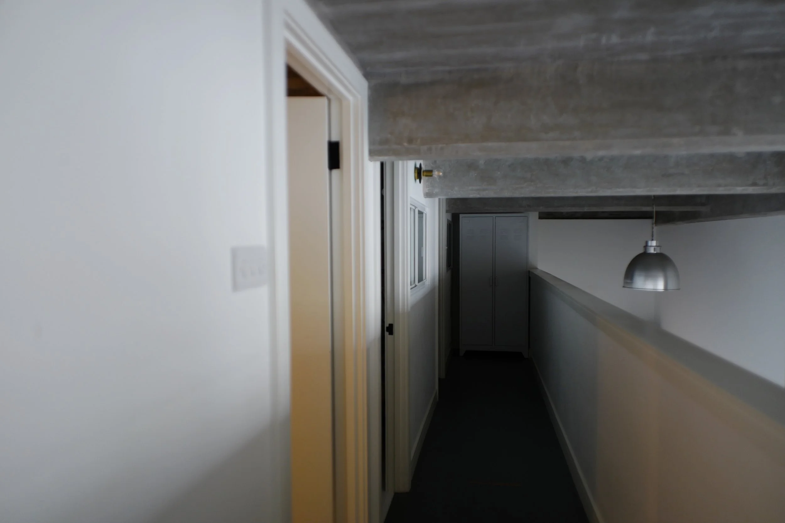 A narrow corridor in a modern interior, with a partially open door on the left, a white wall with a light switch, and a row of cabinets at the end, illuminated by a hanging metallic pendant light.