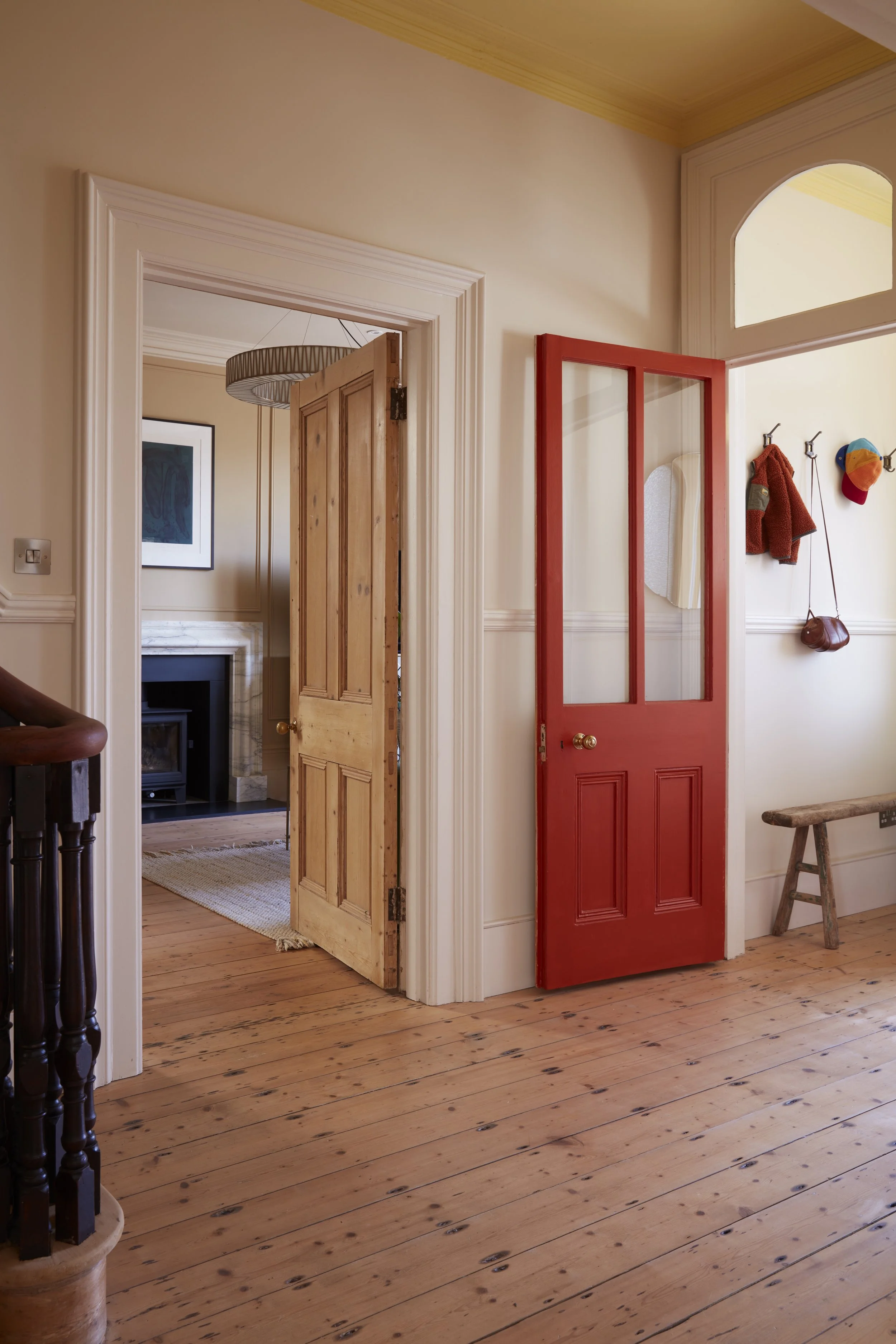 A hallway with wooden floors, white walls, and a yellow ceiling, leading to a room with a fireplace. There is a red door with glass panes on the right, and a wooden bench underneath hanging hooks with a coat, hat, and purse on them.