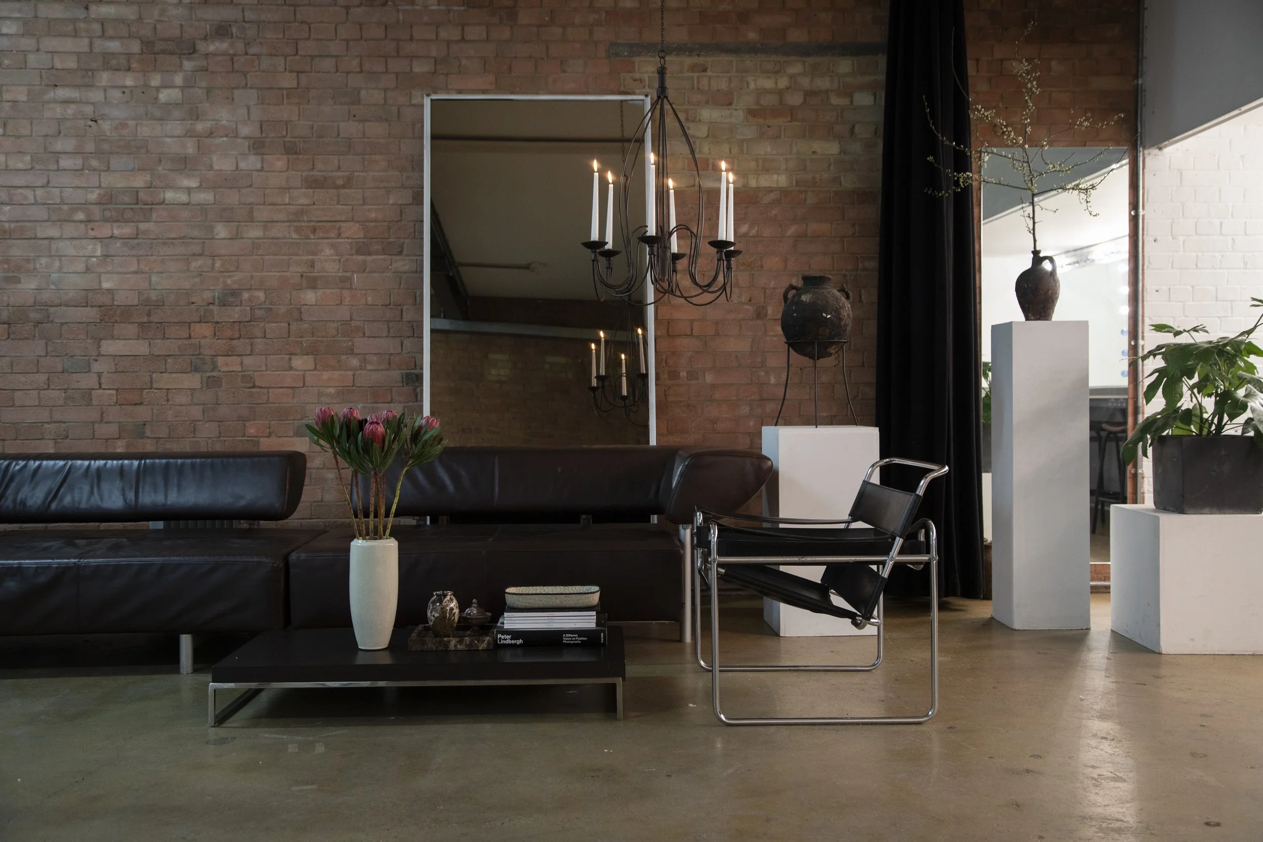 Modern living room with a black leather sofa, a black coffee table with a white vase of pink flowers, a black lounge chair, and decorative pottery on white pedestals, against an exposed brick wall and large mirror.