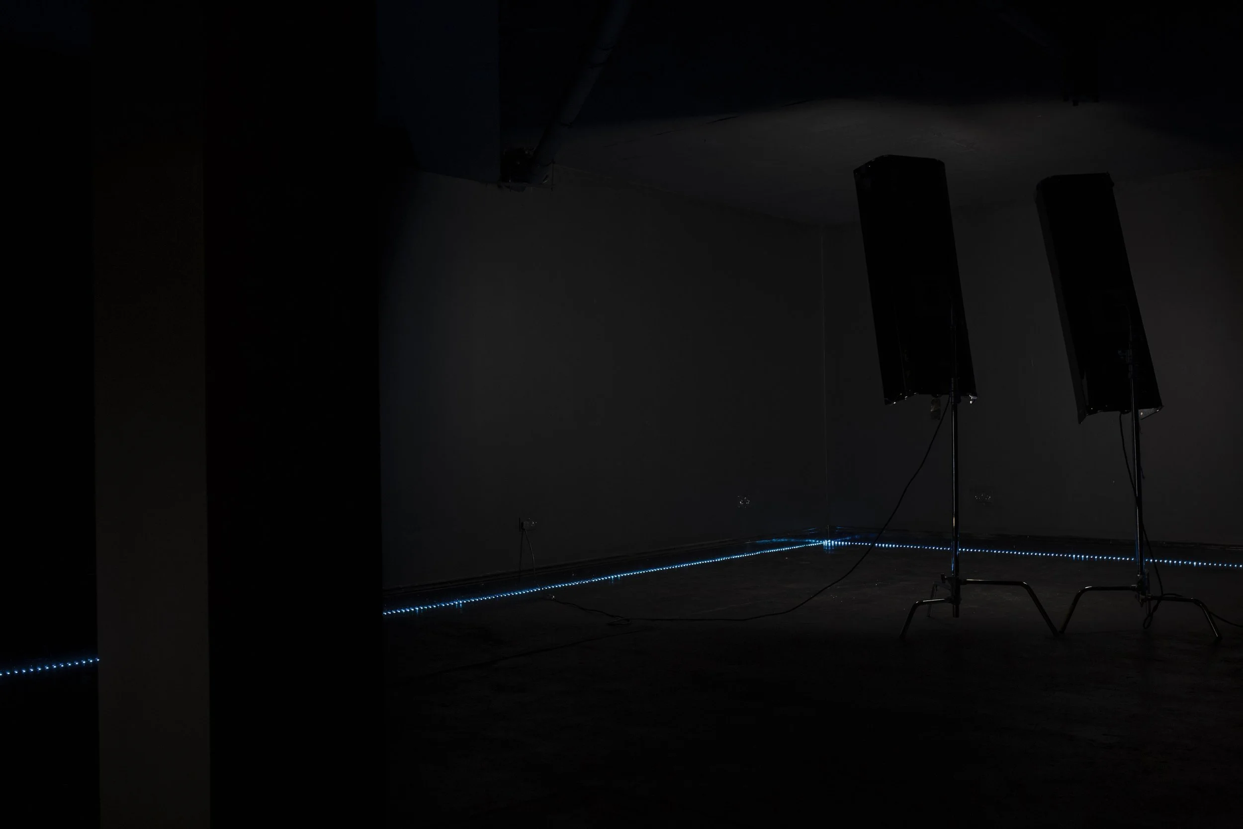 Two tall black speakers on stands in a dark room with a blue LED strip along the base of the wall.