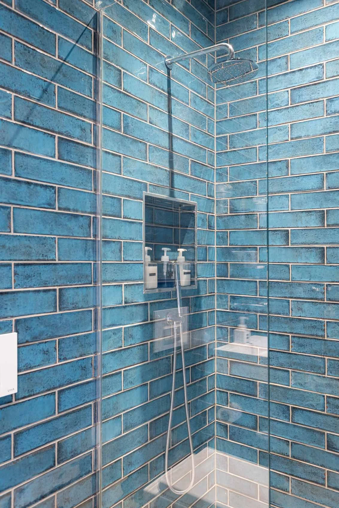 A modern shower with blue subway tiles, a round rainfall showerhead, and a built-in shelf with soap dispensers.