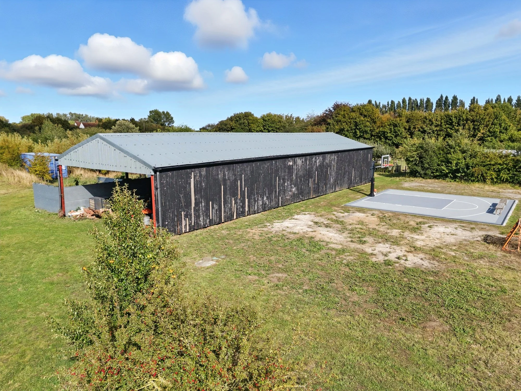 A barn with a corrugated metal roof, surrounded by trees and a sports court in the foreground, under a partly cloudy sky.