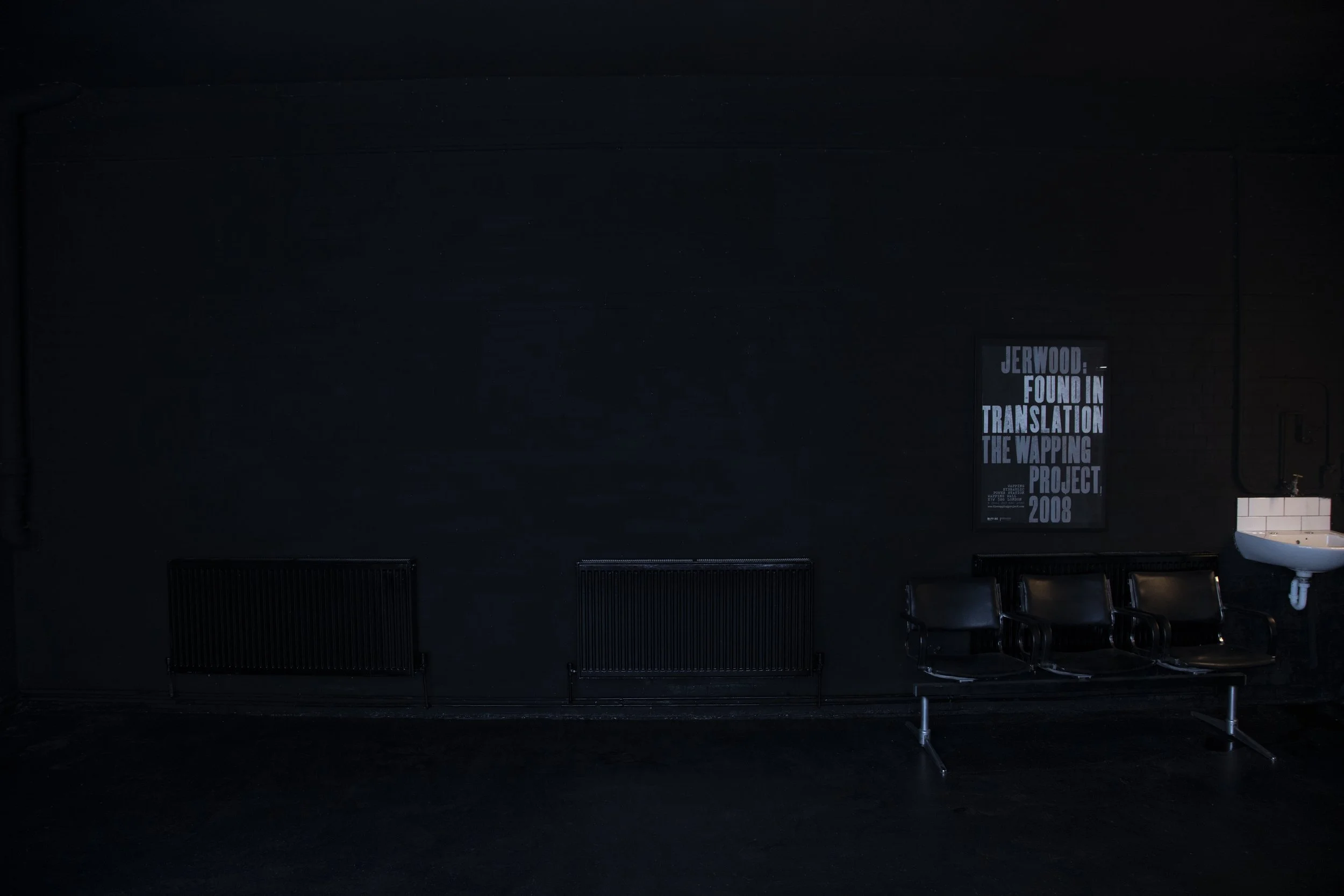 A dark room with black radiators, four black chairs, a framed poster on the wall, and a white sink in the corner.