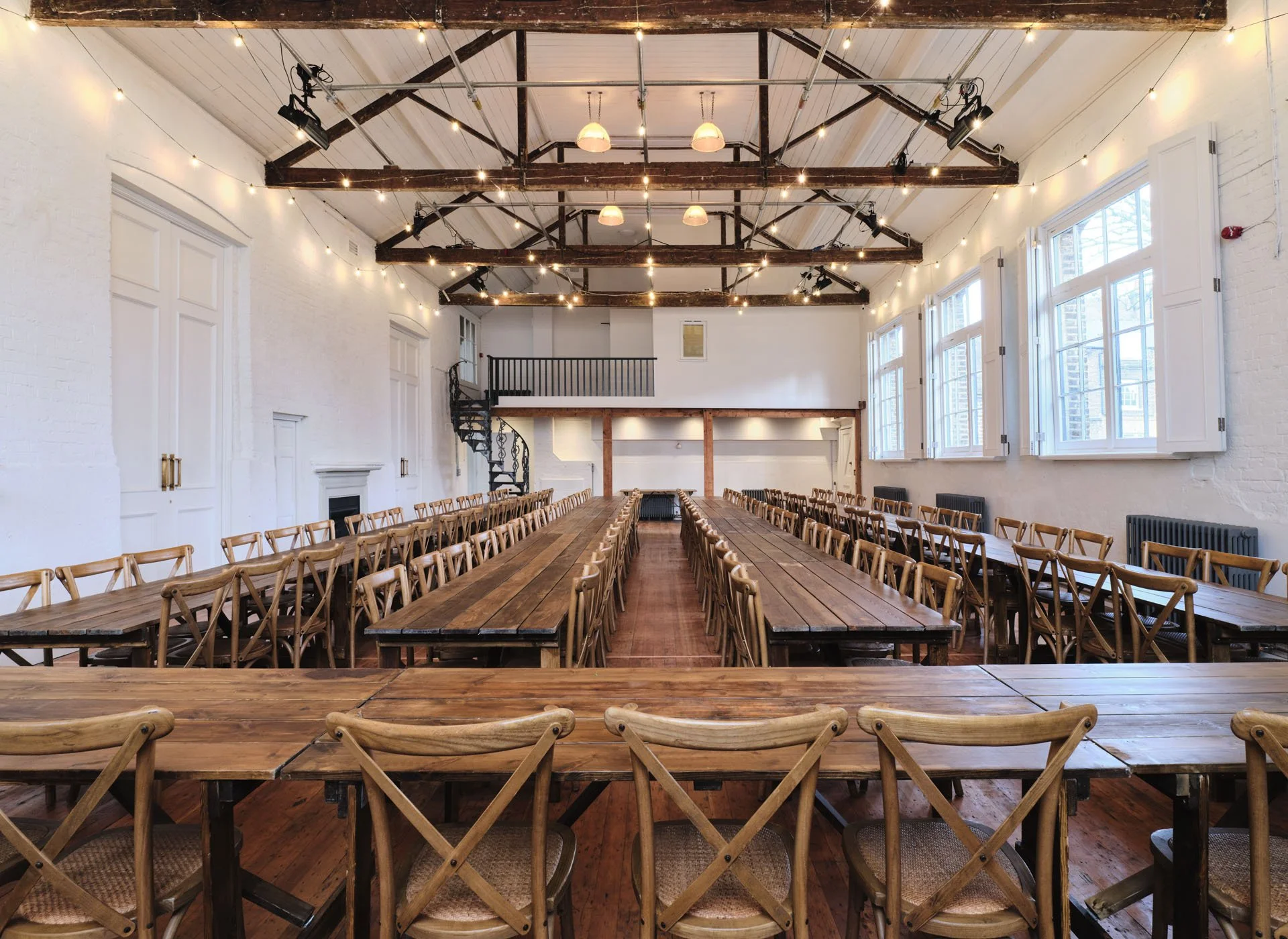 A spacious, bright event hall with wooden tables and chairs arranged in rows, high ceiling with exposed beams and string lights, large windows along the side wall, and a small balcony at the back with a spiral staircase.