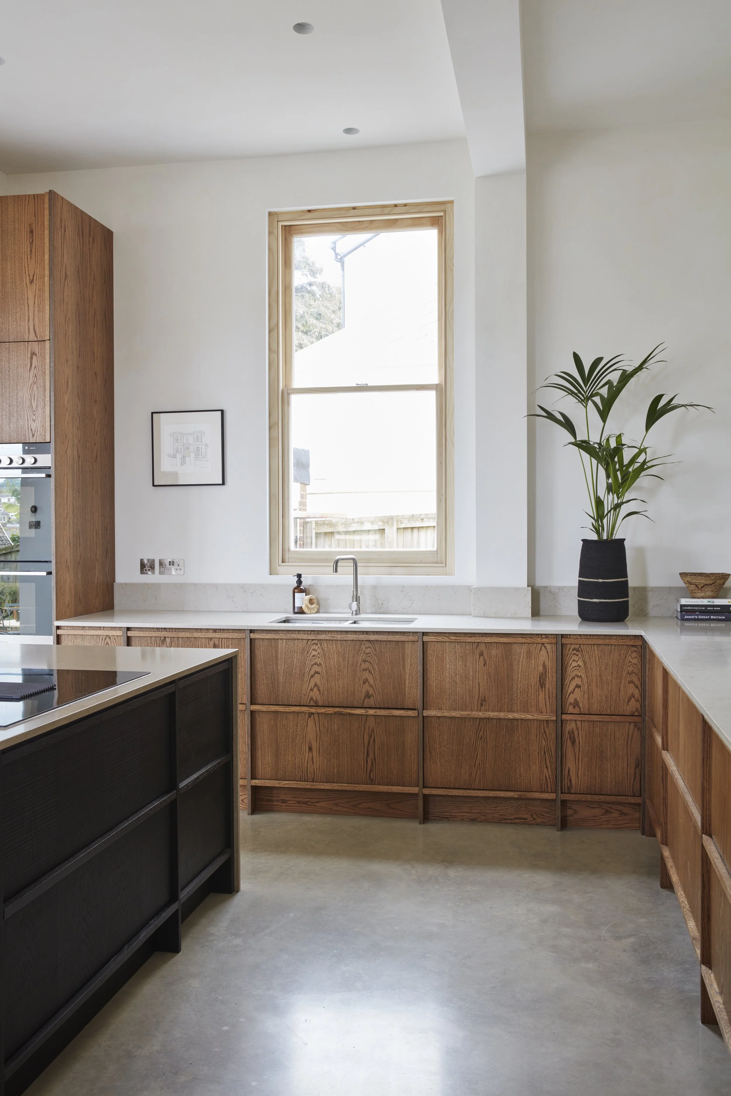A modern kitchen with wooden cabinets, a large window, a black potted plant, and a light-colored countertop.