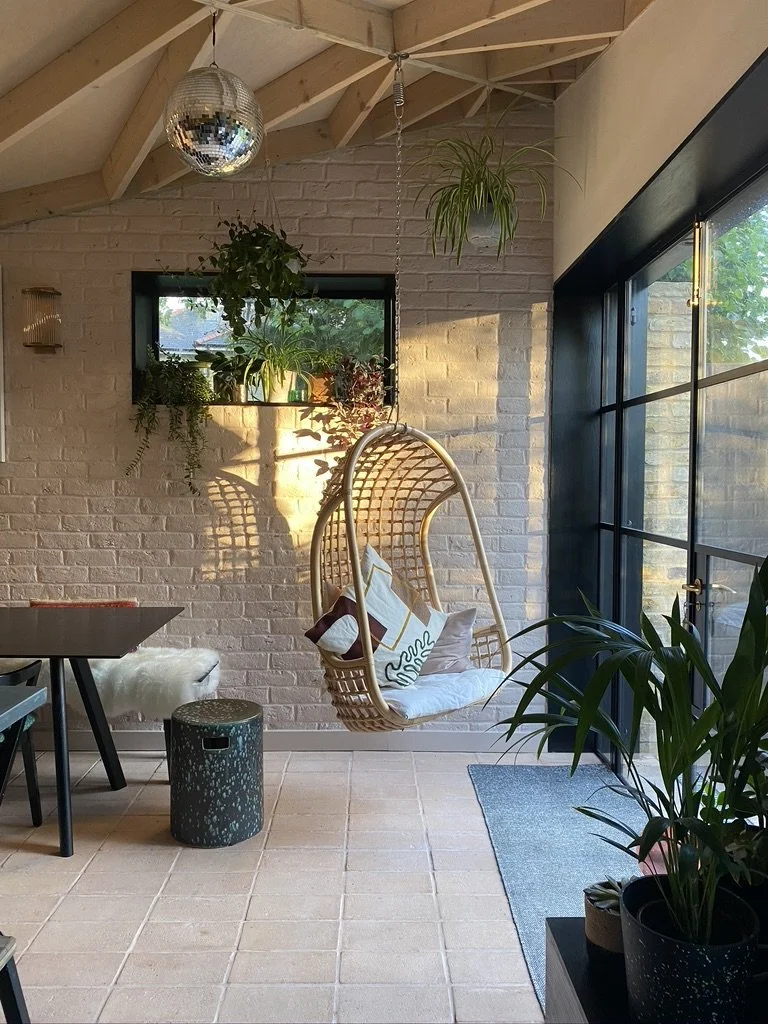 Indoor sunlit sitting area with hanging wicker chair with pillows, potted plants on wall shelf, and large windows revealing outdoor greenery.
