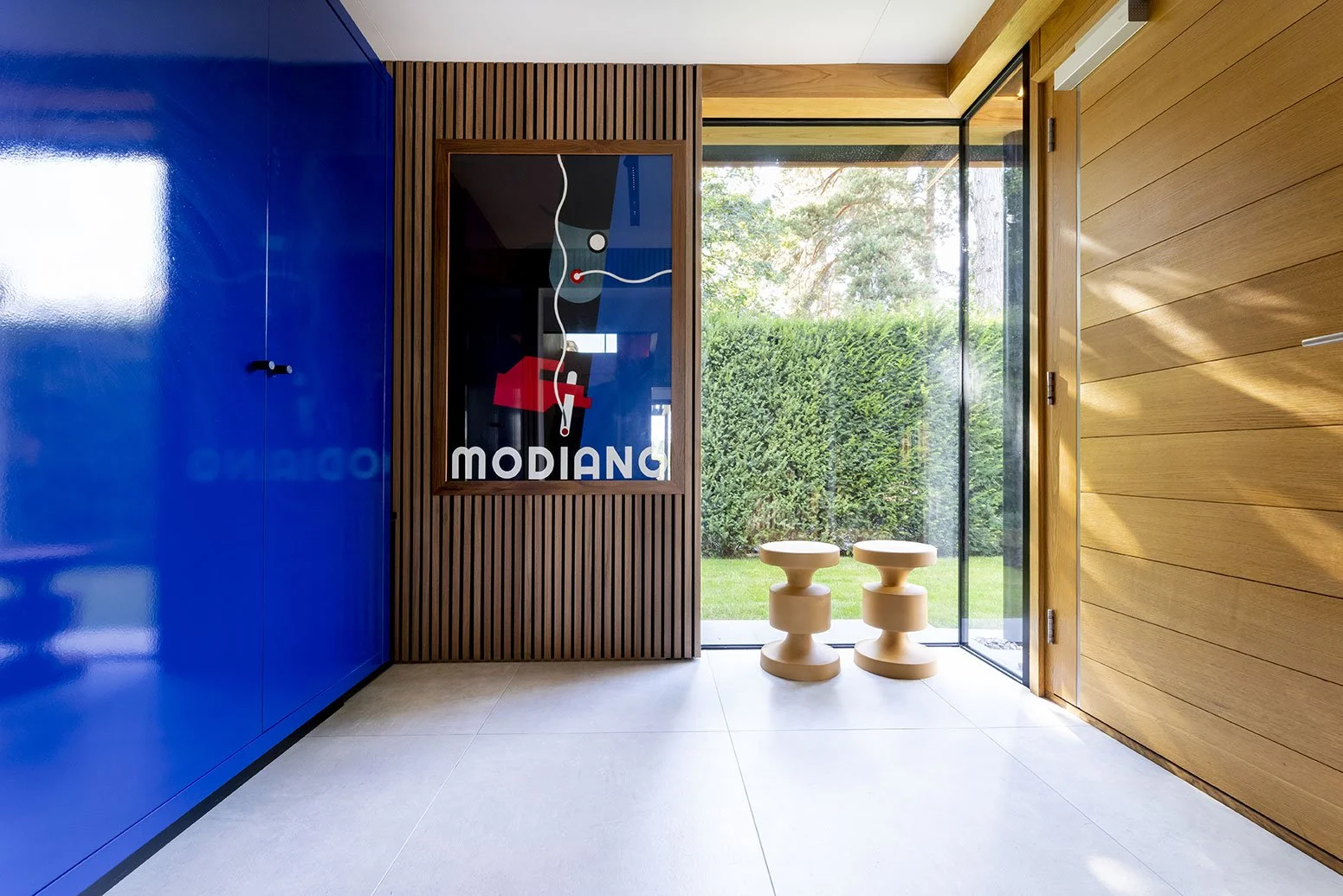 Interior view of a modern room with a blue cabinet on the left, a framed wall art with the word 'MODI' and abstract design, a glass door leading outside to a garden, and two beige sculptural stools near the door.