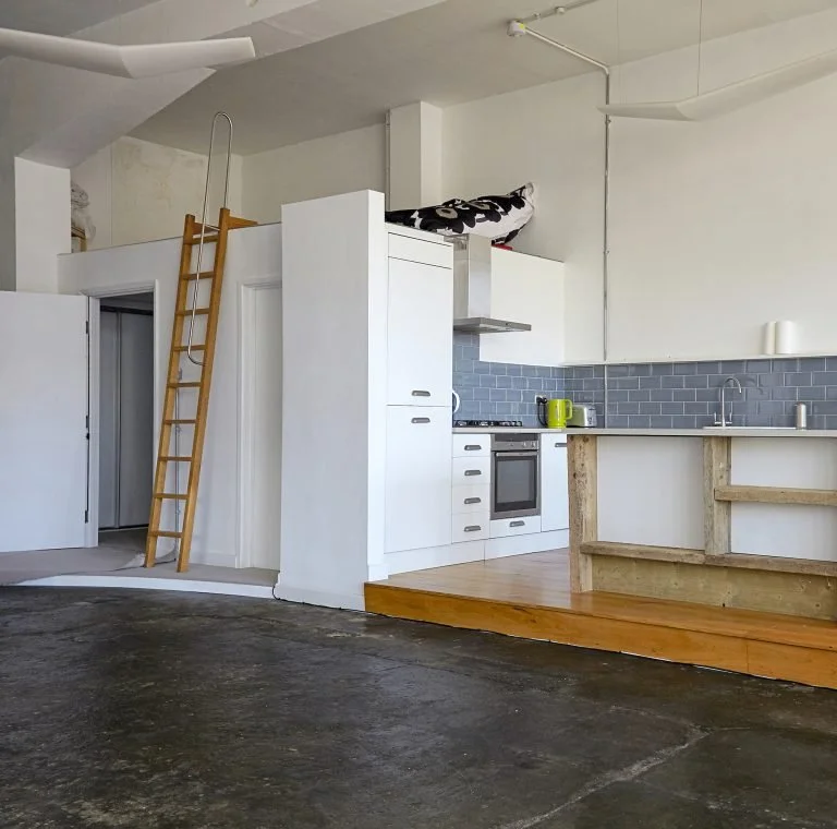 Kitchen with white cabinets, gray tile backsplash, and a small wood platform next to the counter. A wooden ladder leans against the wall leading to an upper loft area.