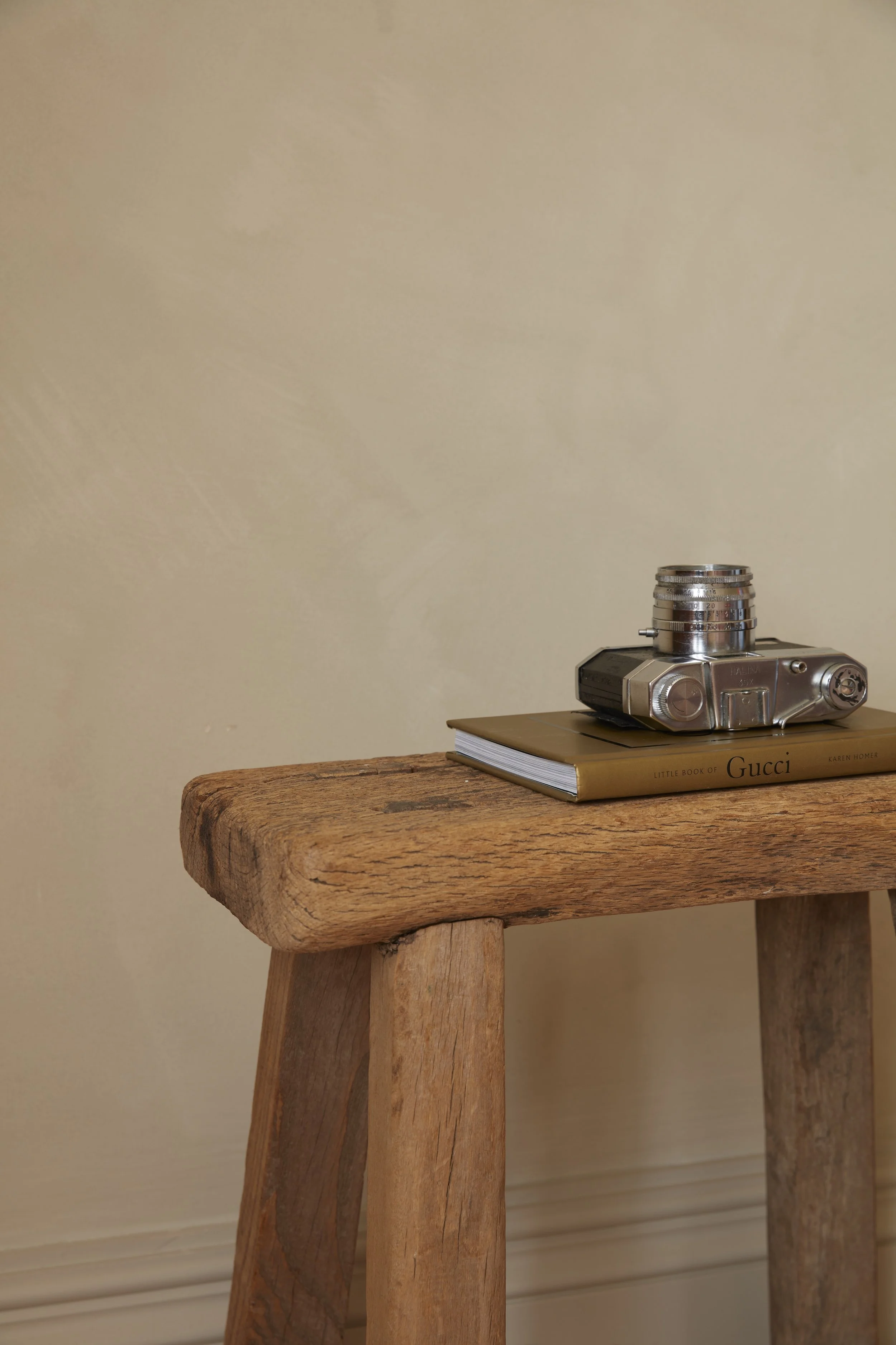 A wooden stool with a vintage camera, a stack of coins, and a book titled 'Little Book of Gucci' on it, against a beige wall.